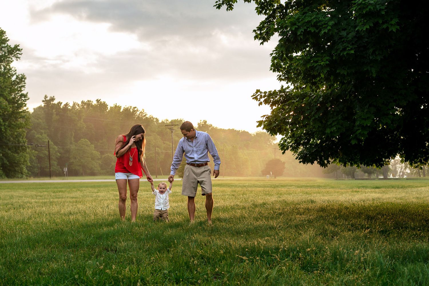 Young family holding toddlers hands while walking through sunlit green meadow at sunset.