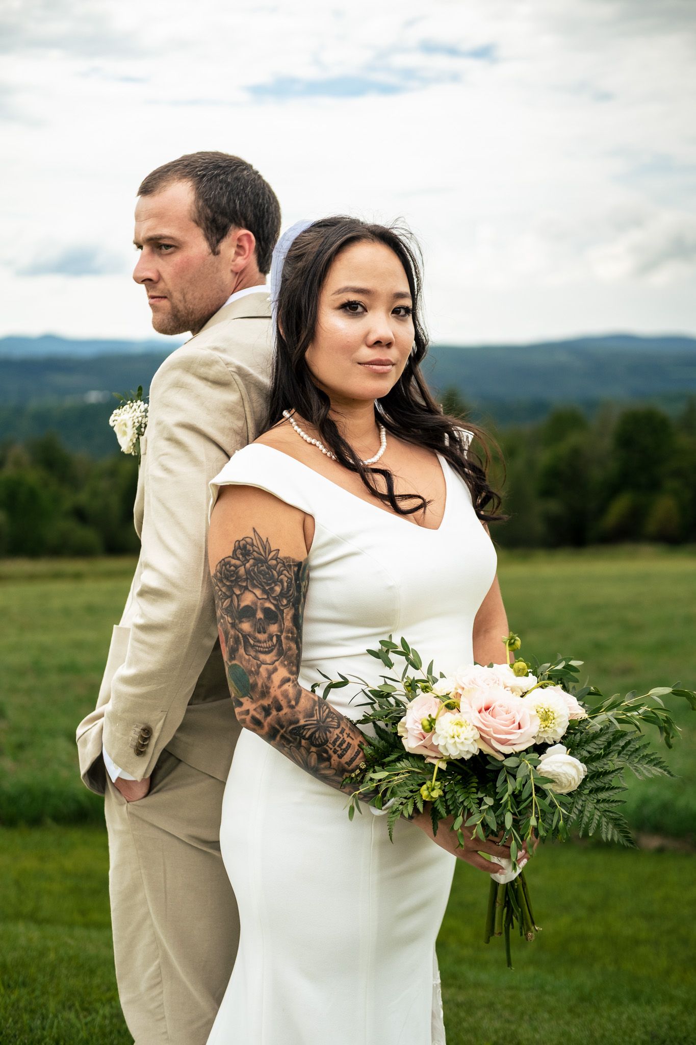 Tattooed bride holds floral bouquet standing back-to-back with groom in scenic mountain field.