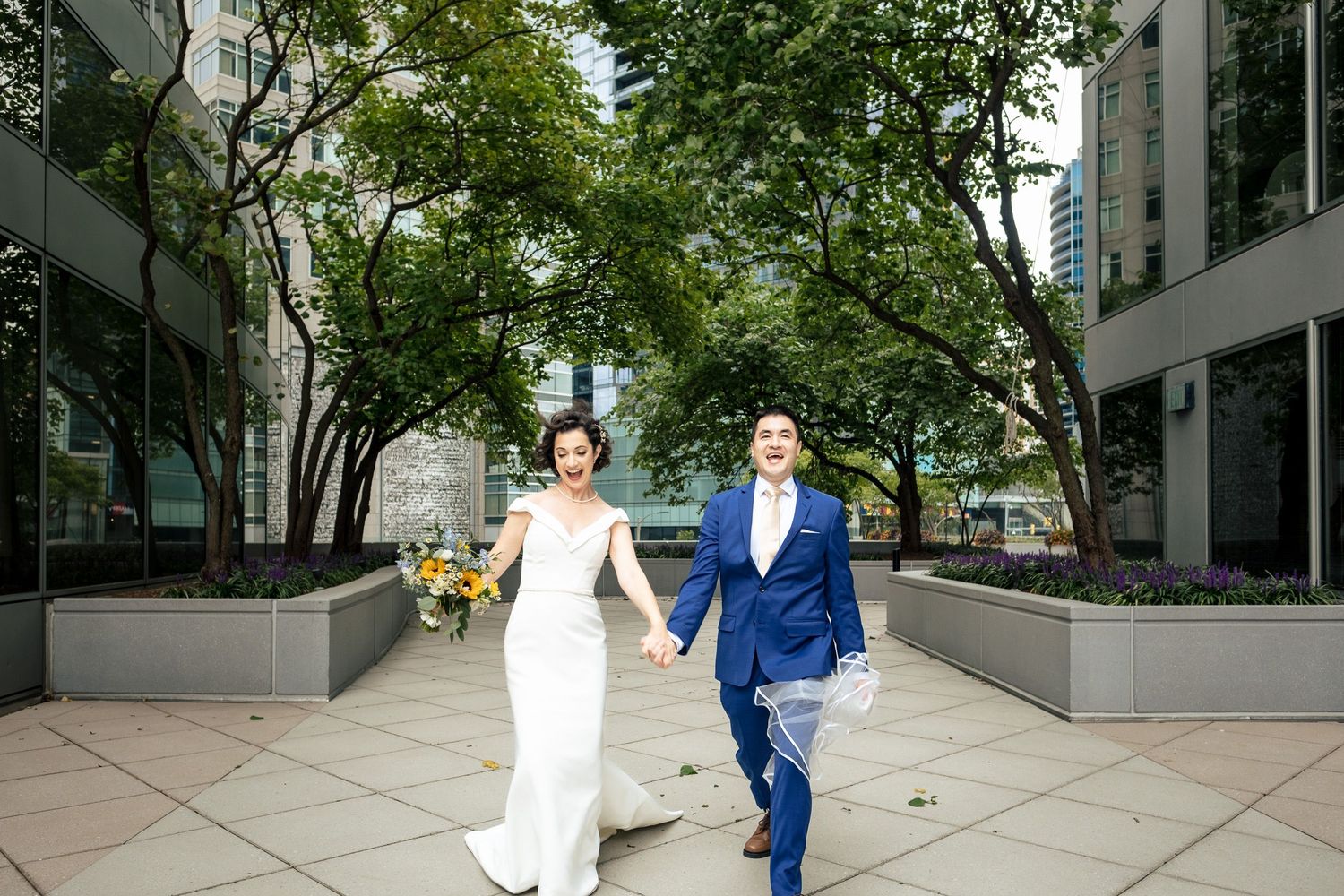 Bride and groom walk hand in hand through a tree-lined urban plaza on their wedding day.