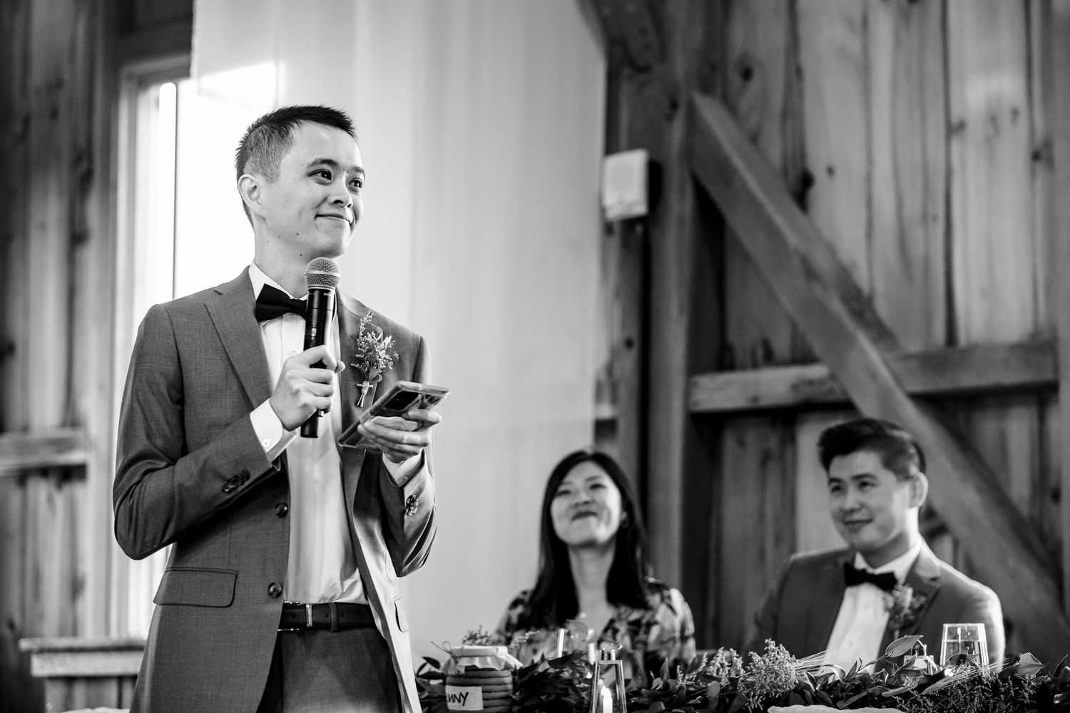 Man in gray suit delivering a wedding toast with microphone at an elegant barn reception.