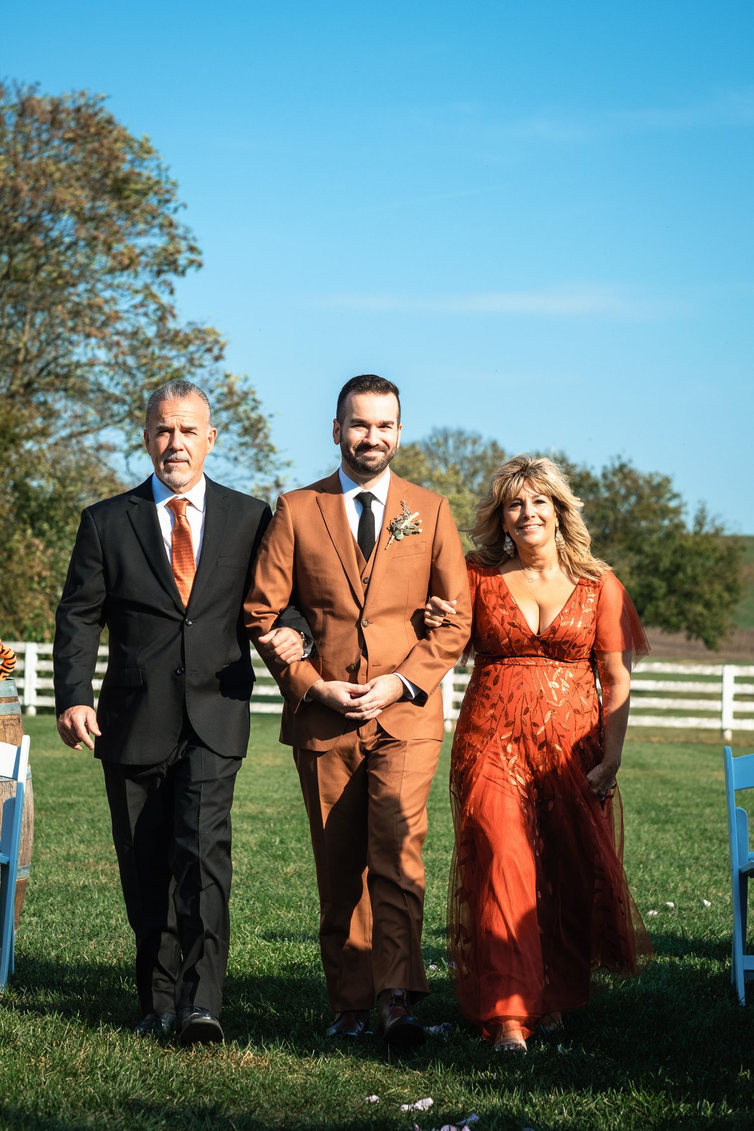 Groom in rust orange suit walks down wedding aisle with his parents at an outdoor farm ceremony.