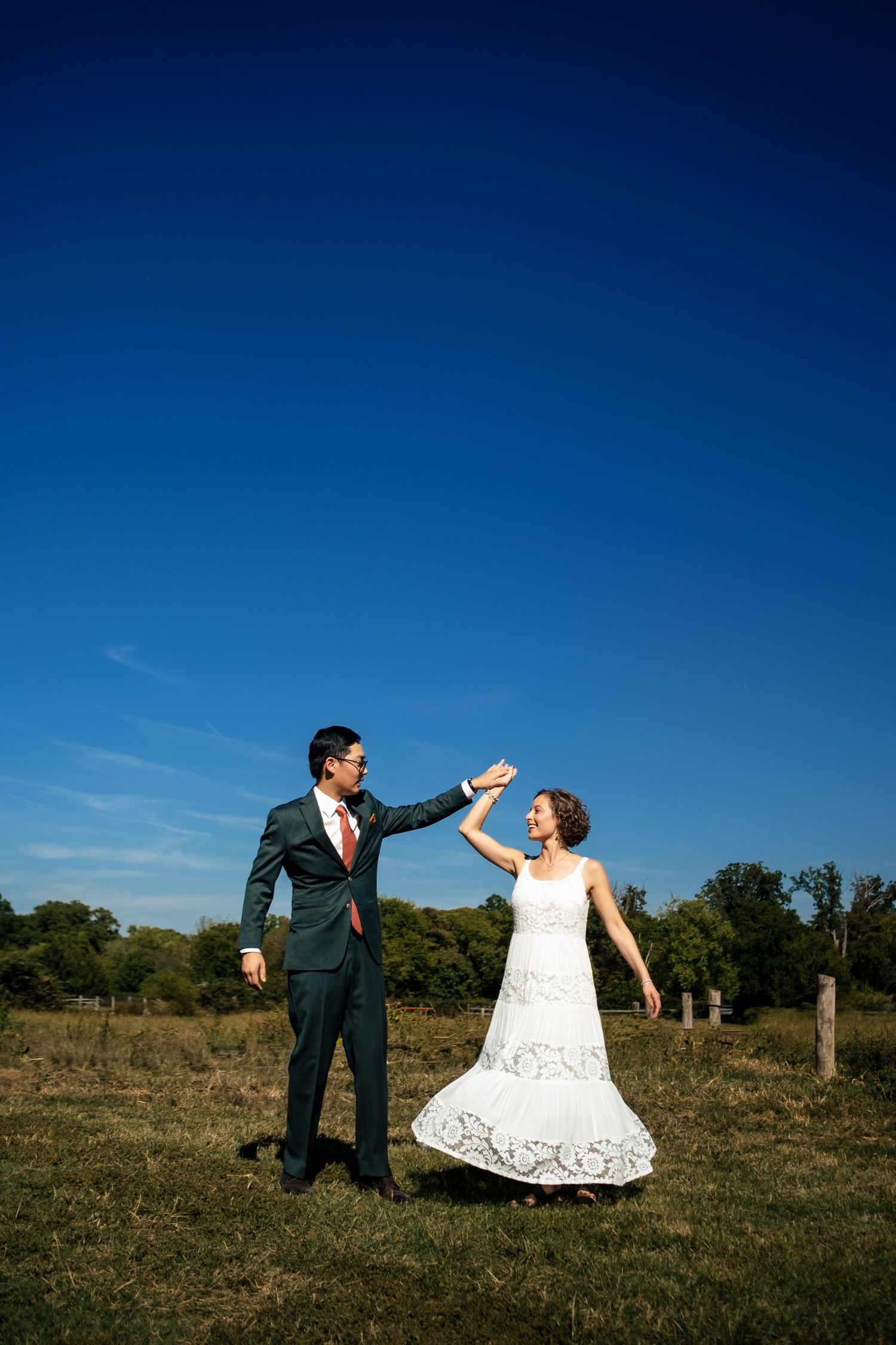 A groom twirls his bride in a lace wedding dress in a sunny open field.