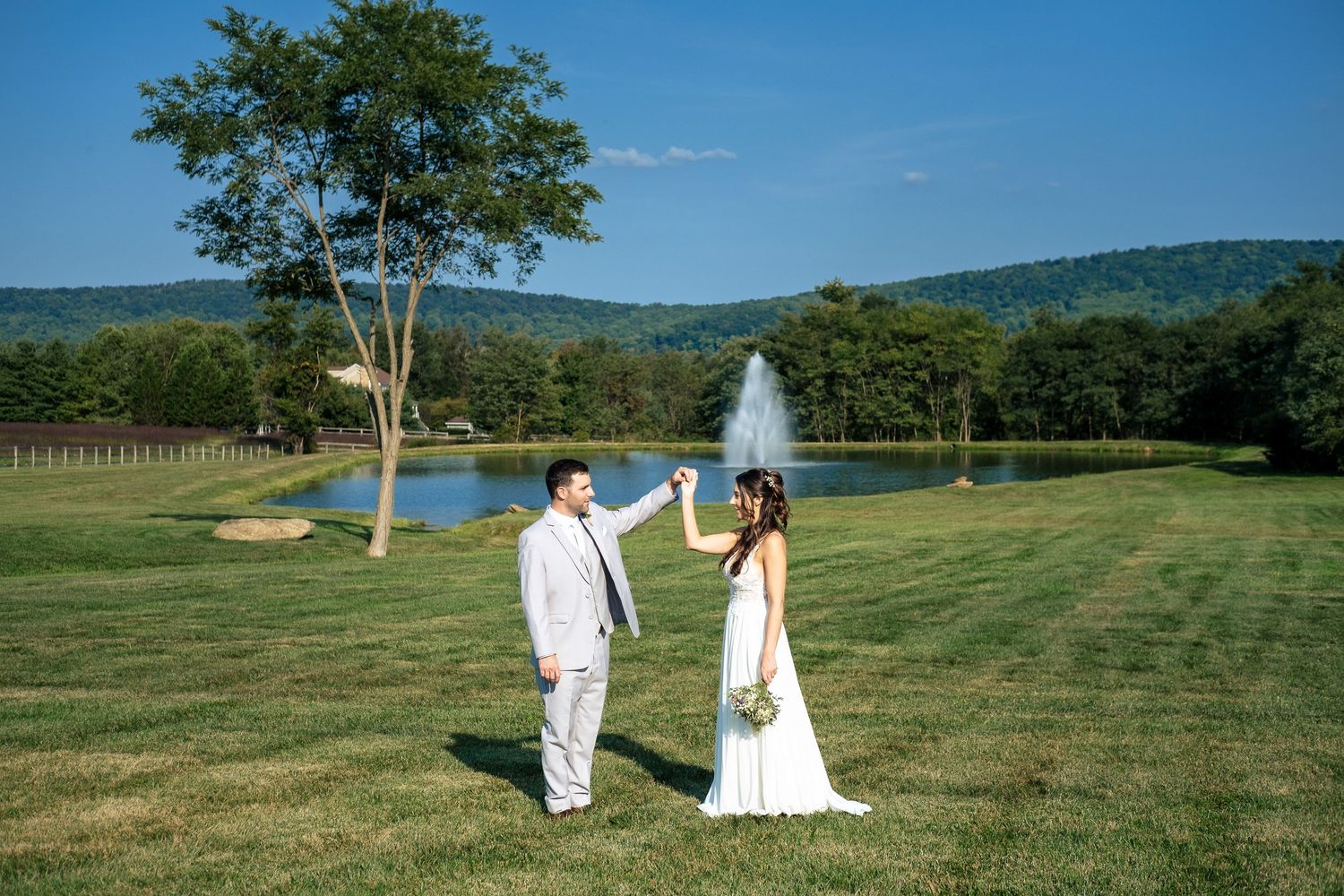 Bride and groom dance on a lush green lawn with a scenic pond and mountain backdrop.