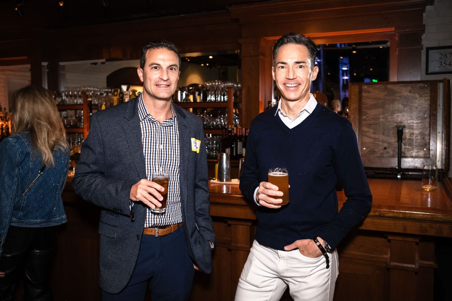 Two men smiling and holding beers at a bar, dressed in smart casual attire at a social networking event.
