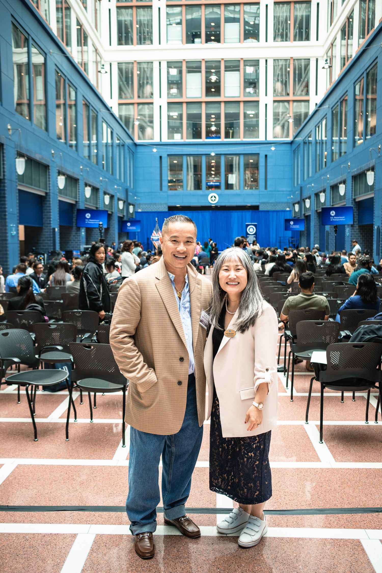Two people smiling together inside a blue-accented university atrium during a graduation ceremony.