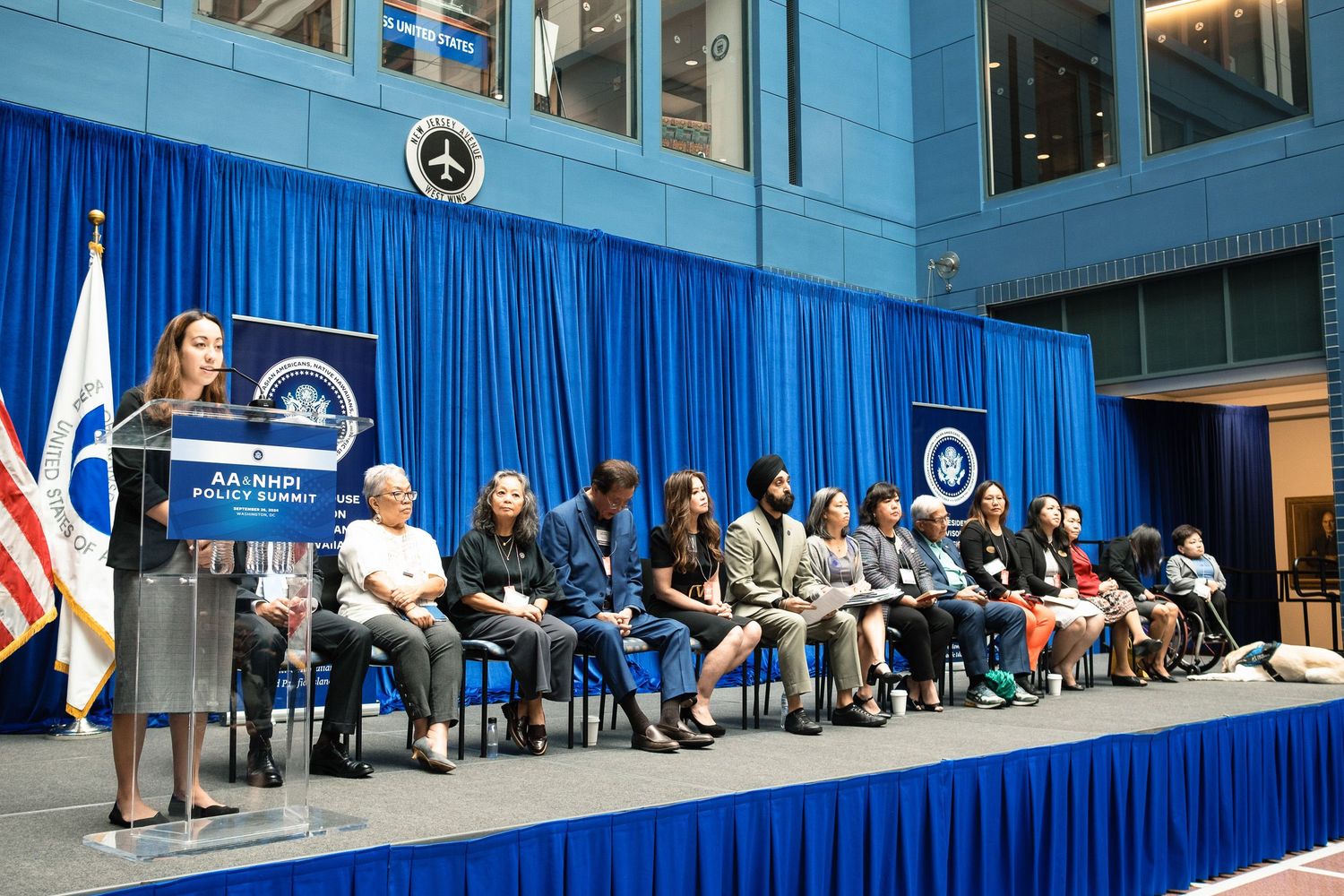 A speaker addresses a diverse panel of women seated on a blue-draped stage at a formal government ceremony.