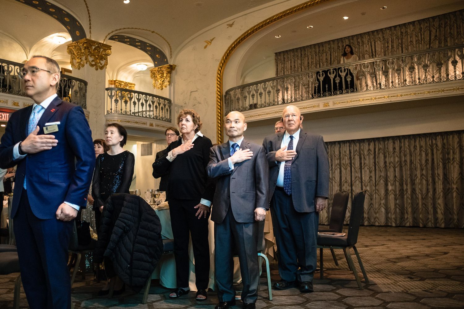 Group of elegantly dressed people standing with hands over hearts during a formal ceremony in a grand ballroom venue.