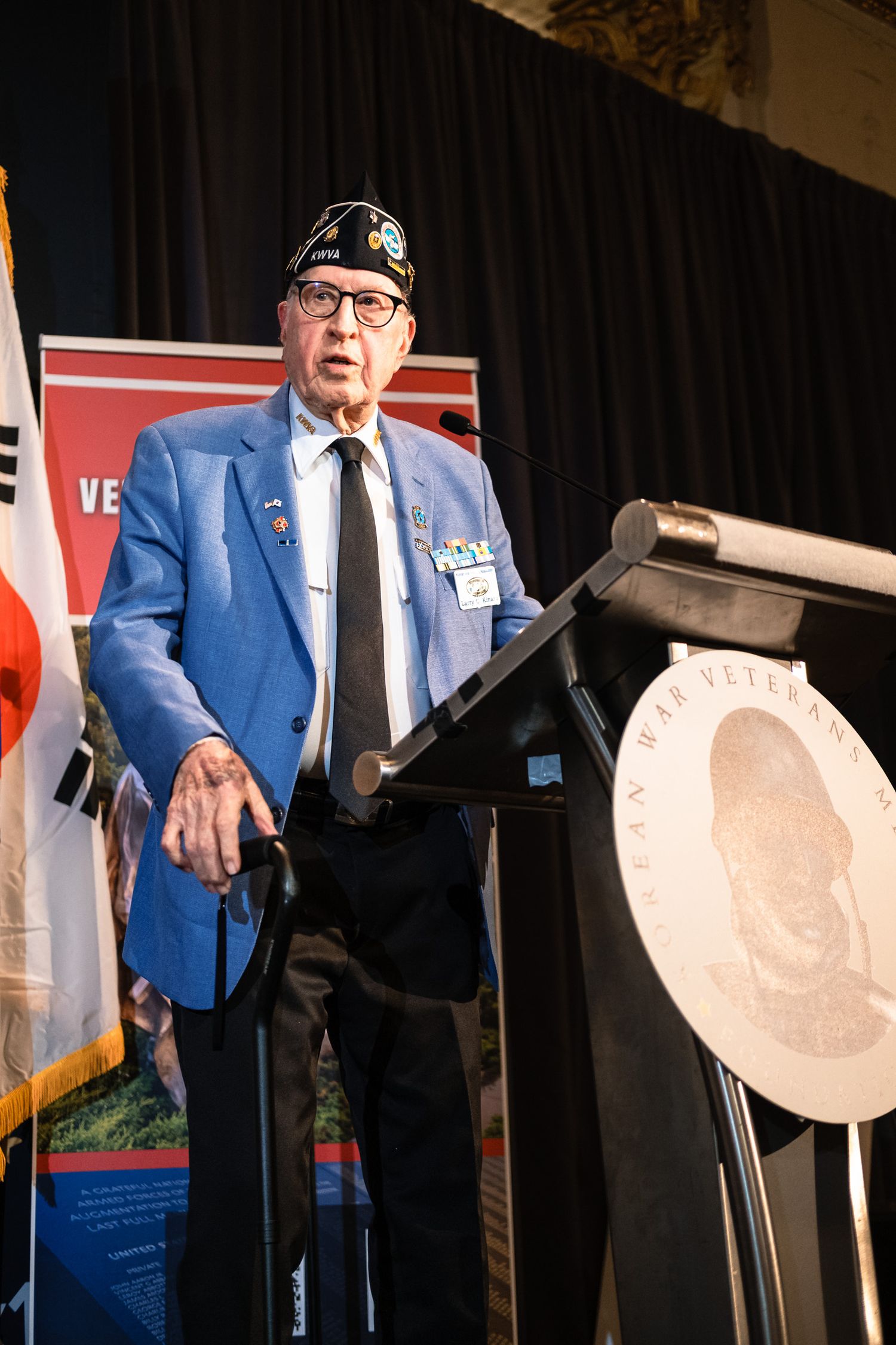 Korean War veteran in blue blazer and military cap speaks at podium during veterans memorial foundation ceremony.
