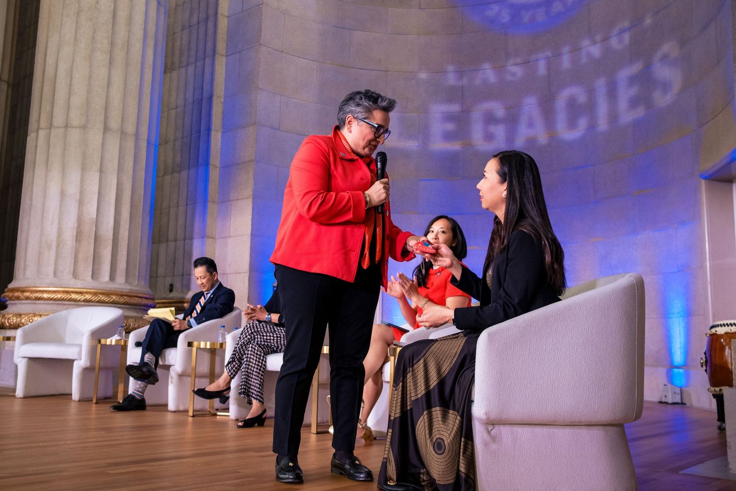 Speaker in red jacket interviews seated woman at Lasting Legacies event on elegant stage.