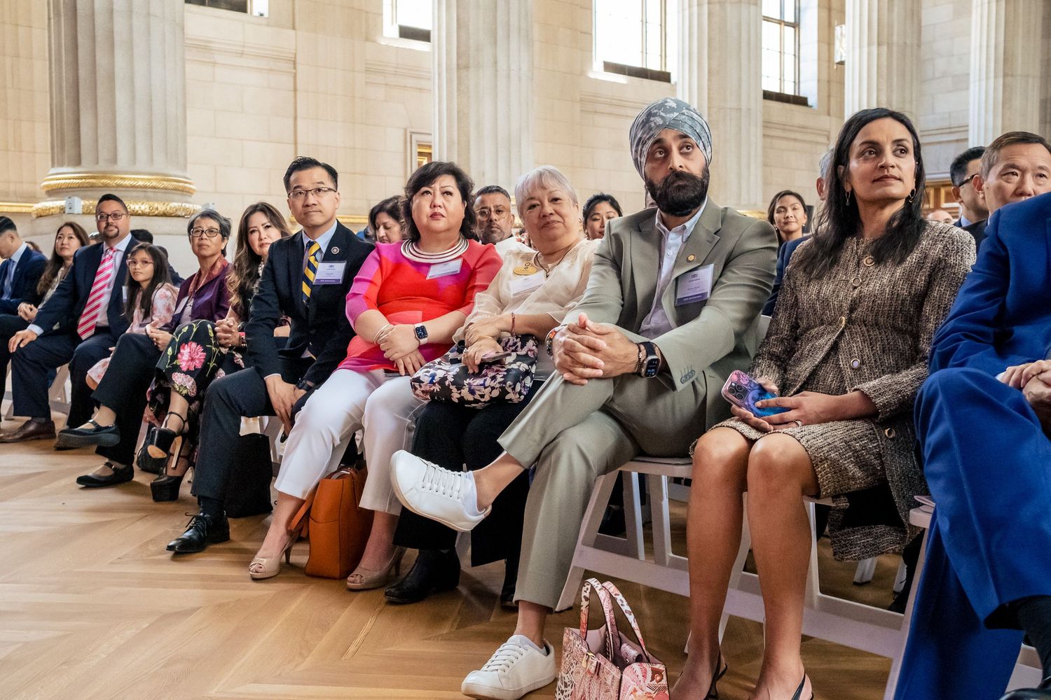 Diverse group of elegantly dressed guests seated at a formal indoor ceremony in a grand classical building.