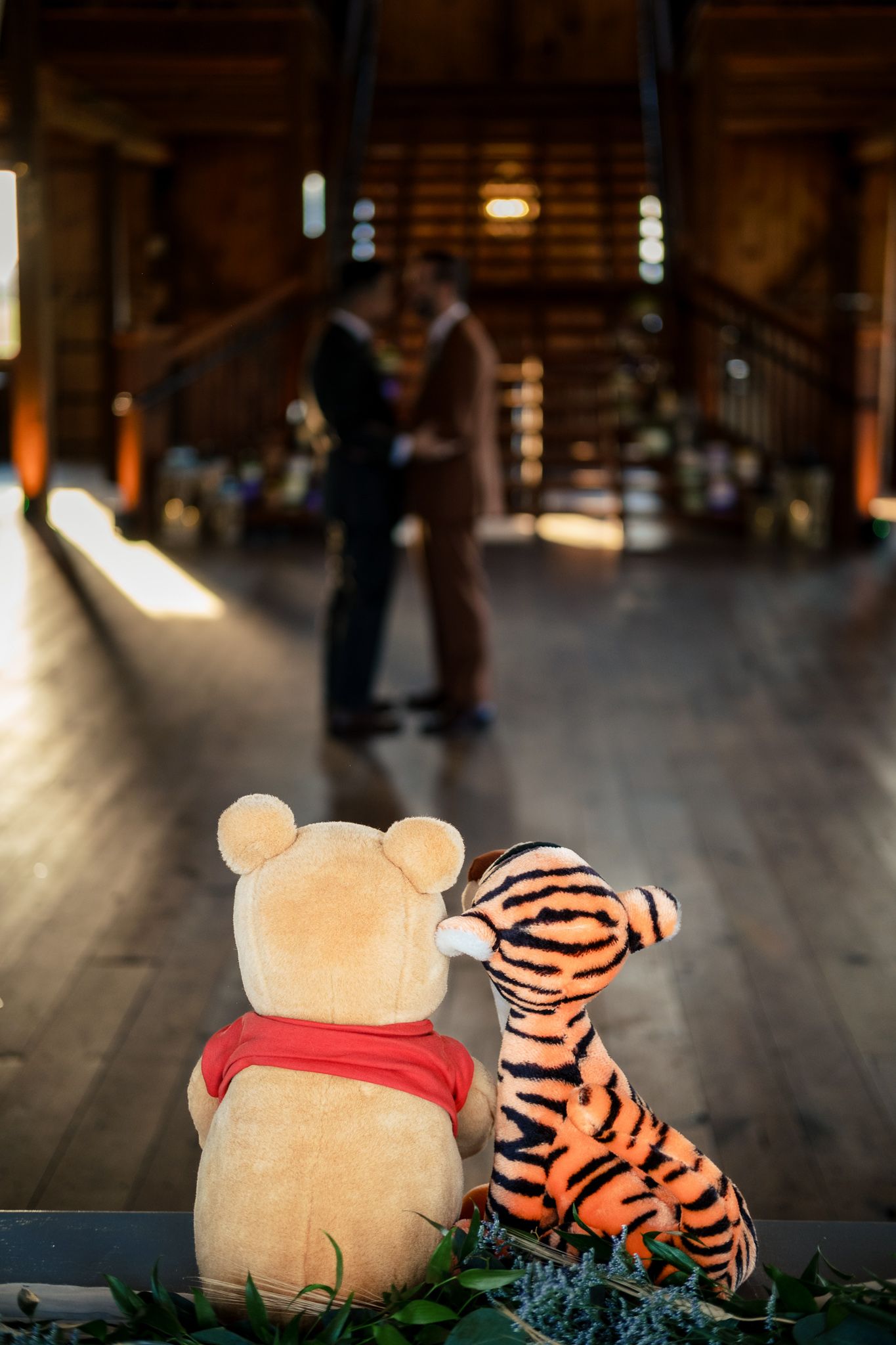 Winnie the Pooh and Tigger plush toys watching couple embrace in rustic barn wedding venue.