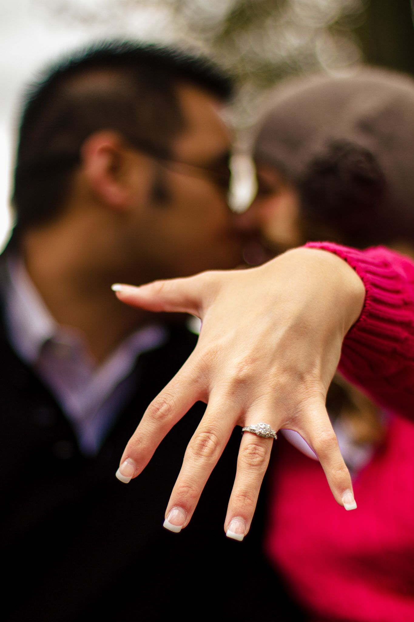 Engaged couple kissing while woman displays diamond engagement ring close to camera outdoors.