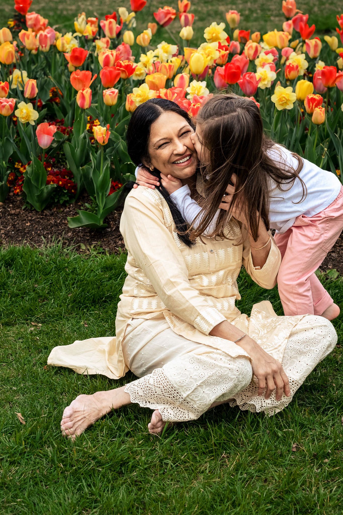 Woman in yellow dress laughing joyfully as young girl hugs and kisses her cheek in tulip garden.