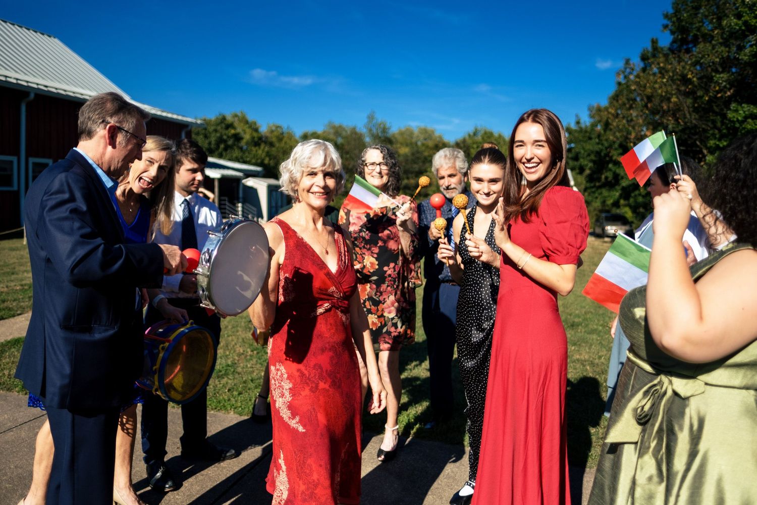 Joyful wedding guests in red dresses wave Italian flags and maracas during a festive outdoor celebration.