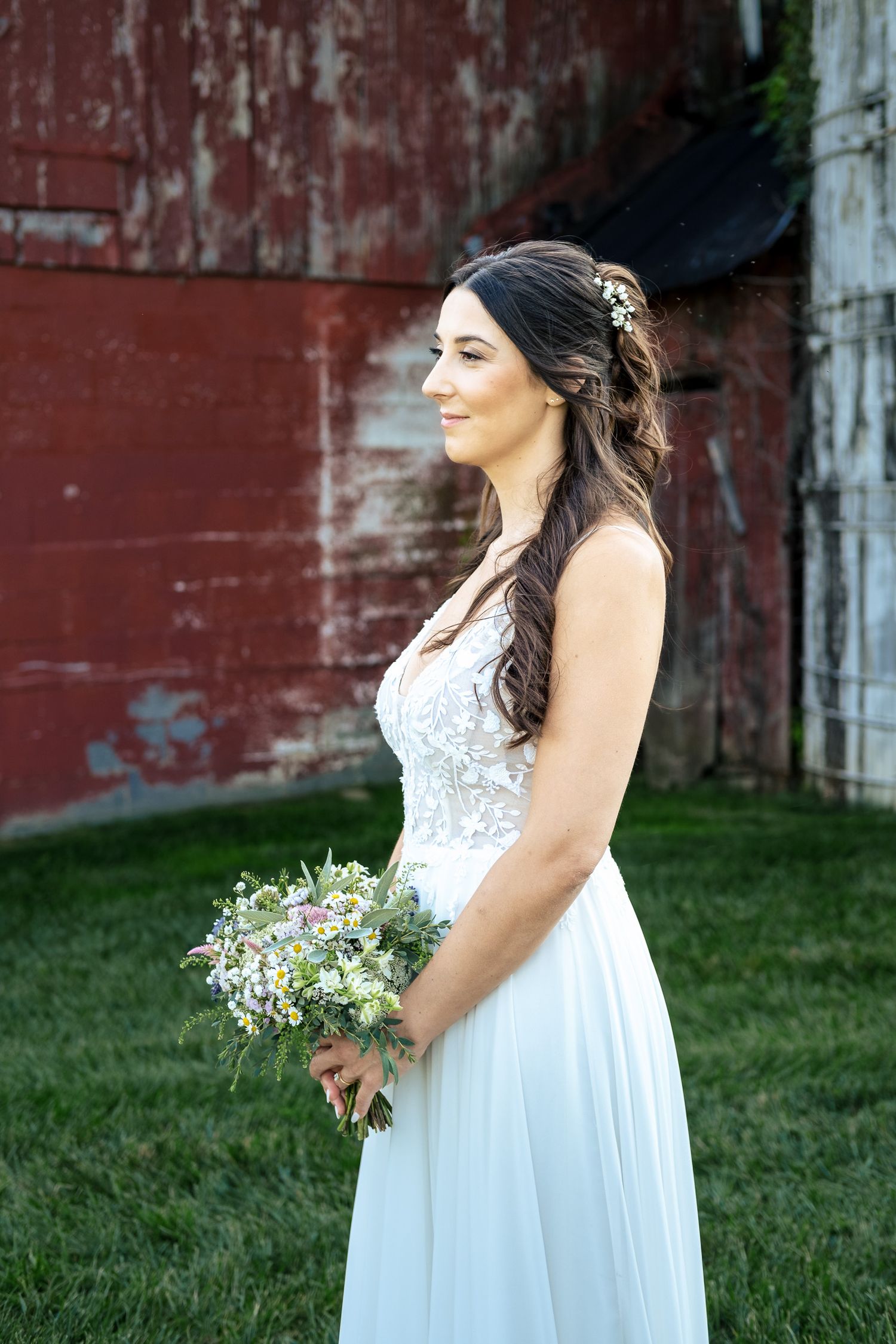 Smiling bride in white lace gown holds a wildflower bouquet standing beside a rustic red barn.