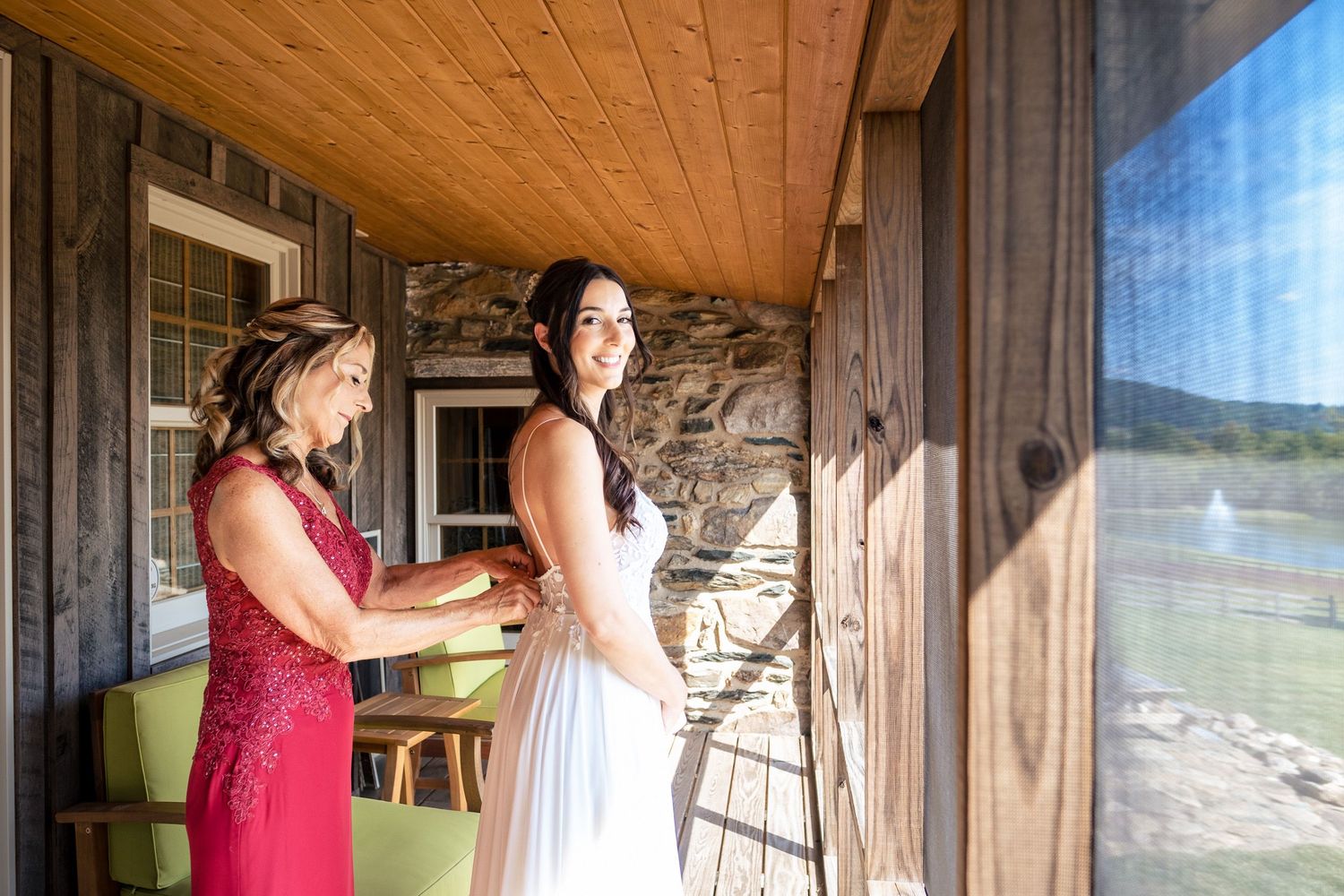 Mother helping bride in white wedding gown on a sunlit porch with scenic lake views.