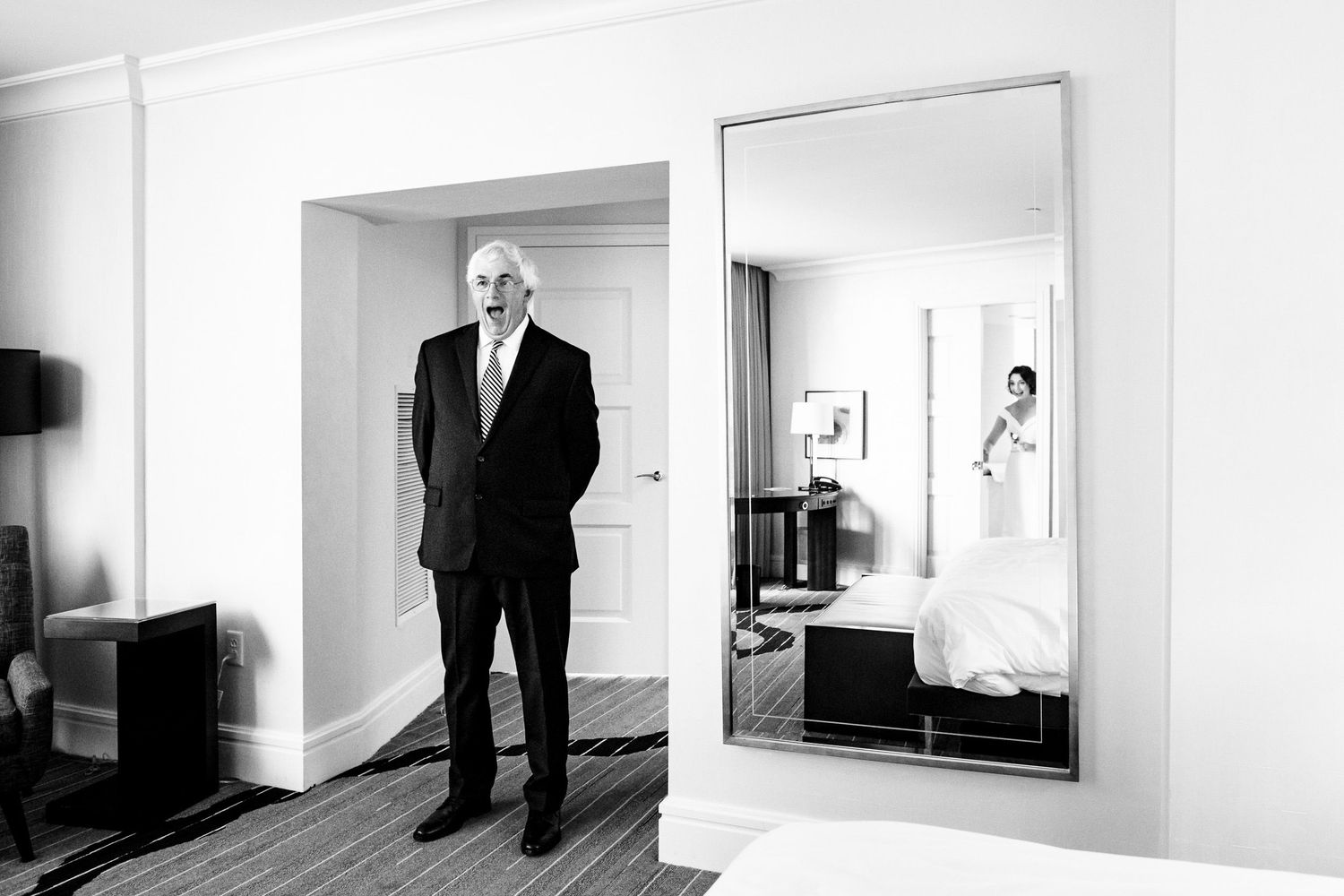 Elegant black and white photo of older man in suit standing in hotel room doorway with bride reflected in mirror.