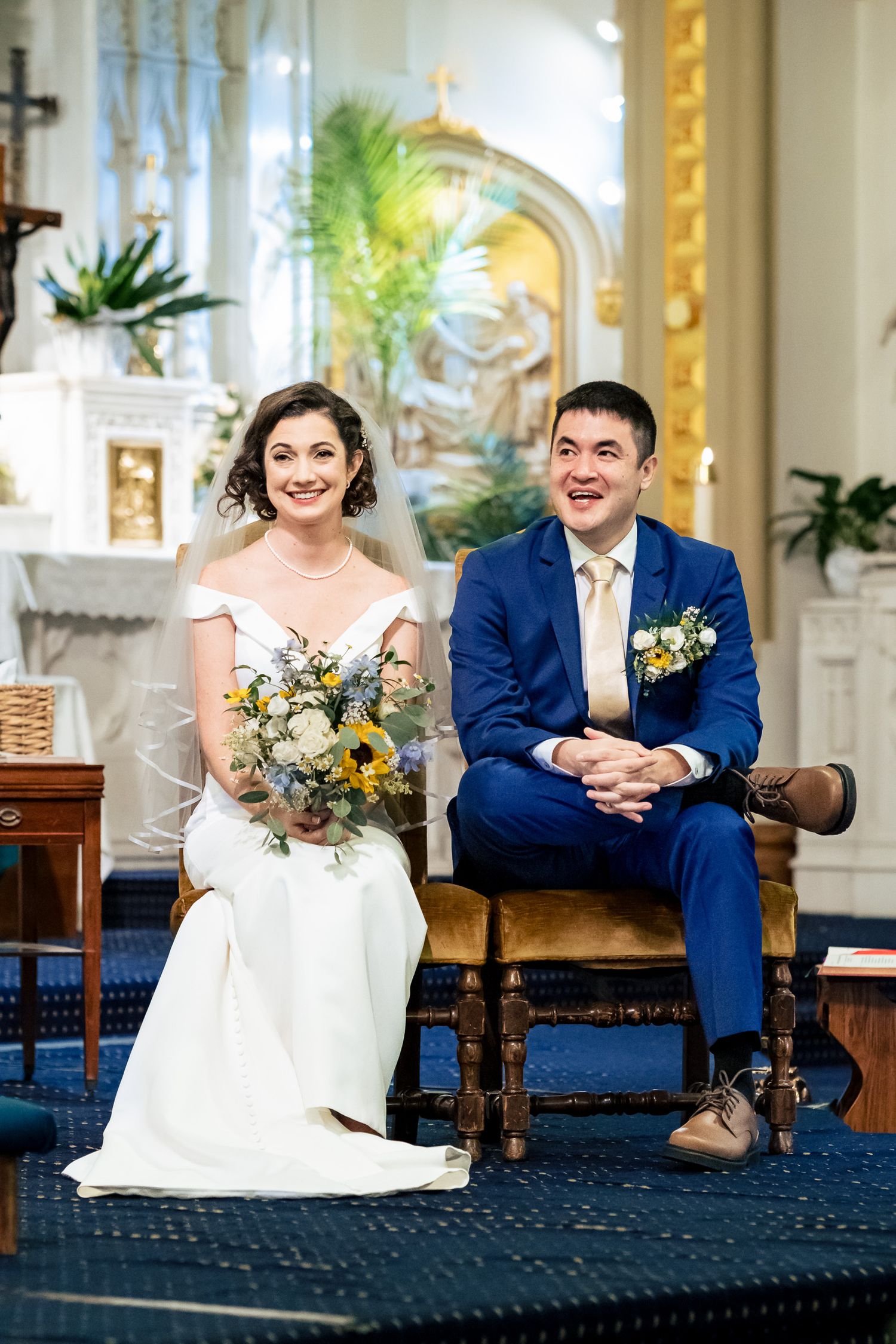 Bride and groom sit together smiling at the altar during their Catholic church wedding ceremony.