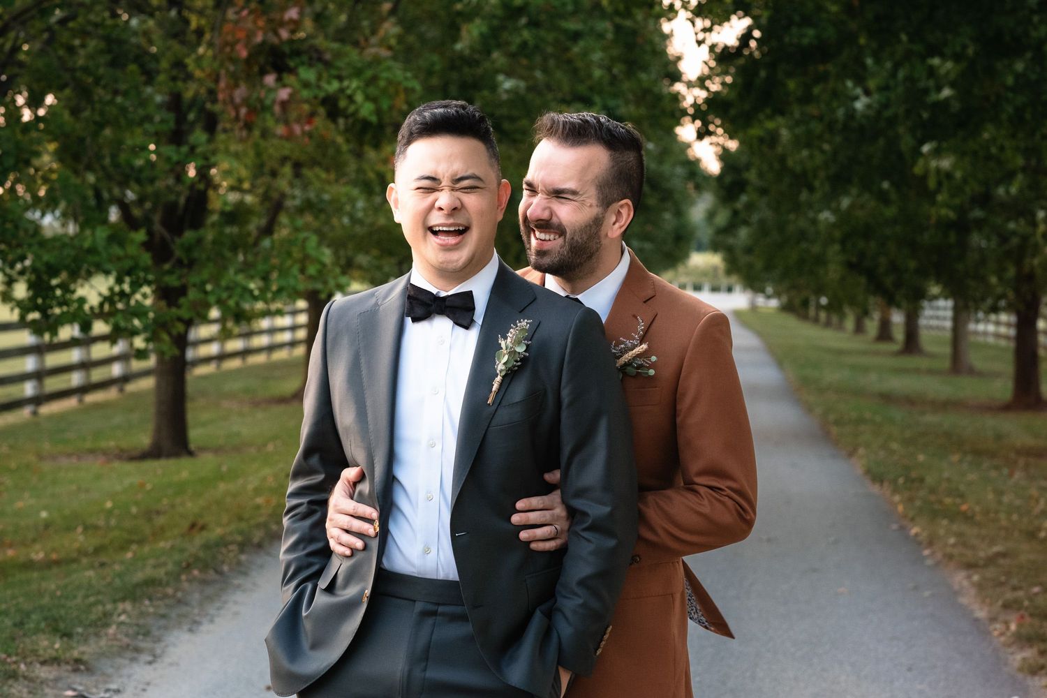 Two grooms walking and laughing together on a tree-lined path at their outdoor wedding.