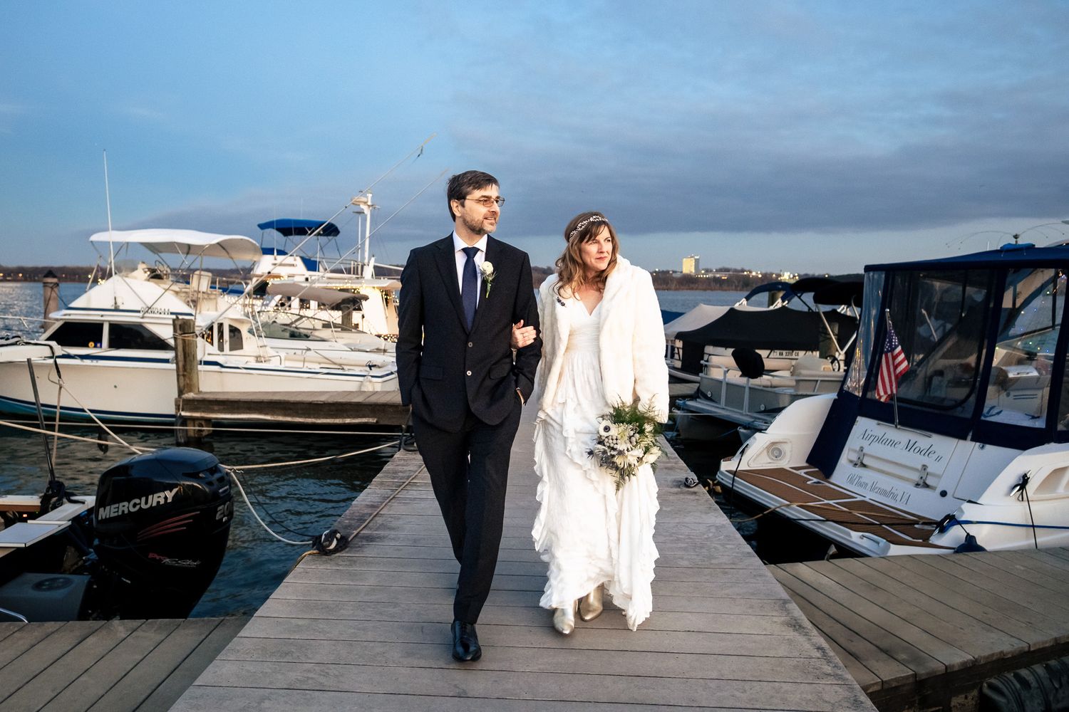 Bride and groom walk hand in hand along a marina dock with boats in the background.