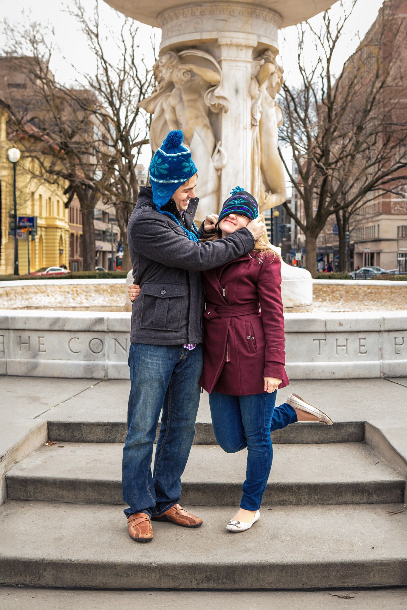 Couple in matching blue winter hats embrace playfully near a stone fountain in a city park.