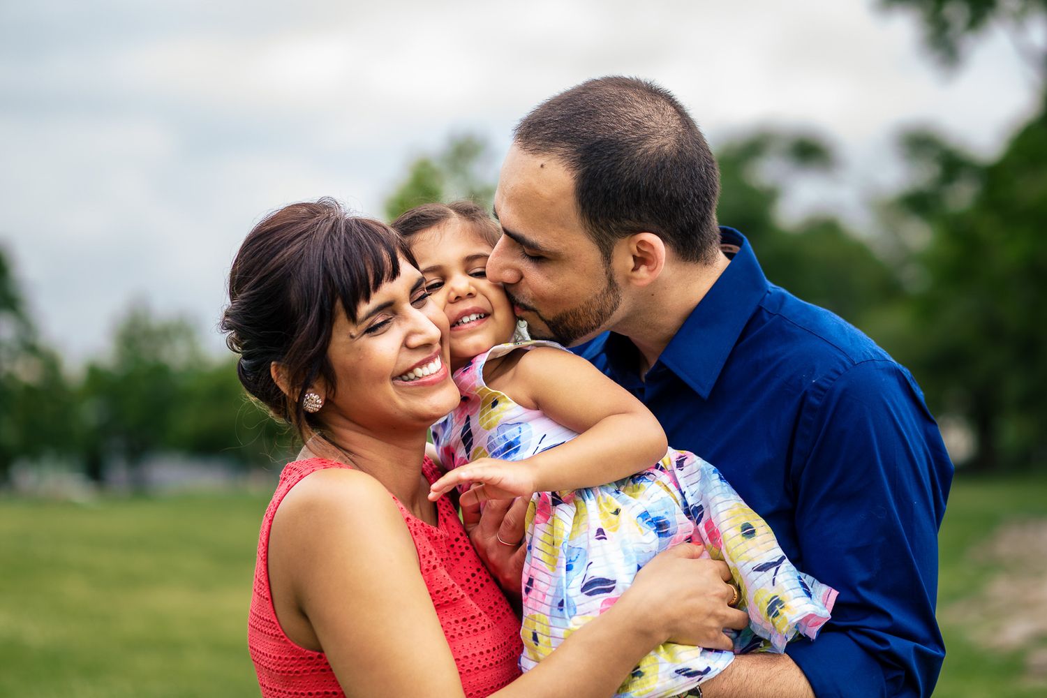 Parents kissing and hugging their laughing young daughter in outdoor family portrait.