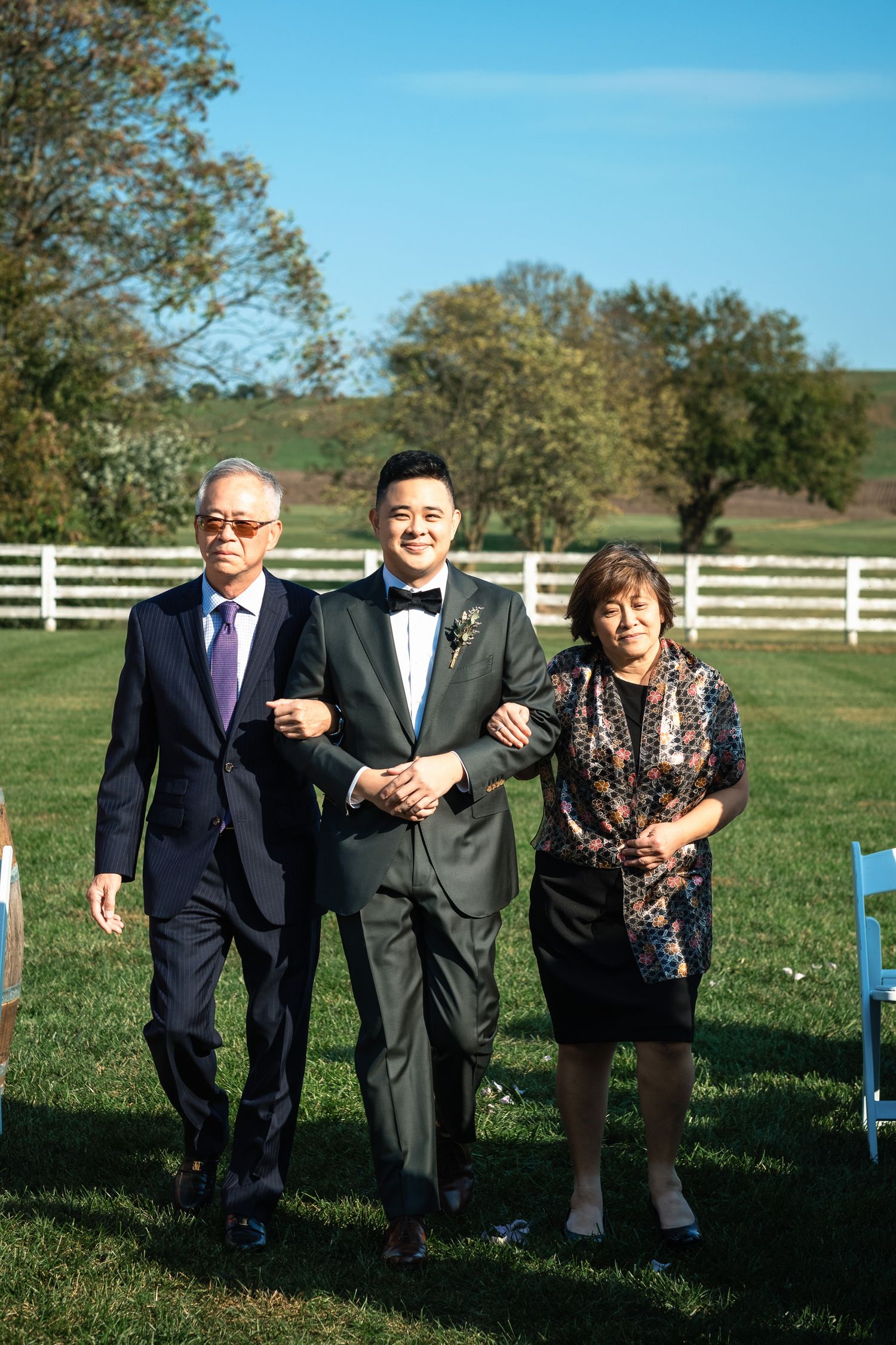 Groom in black tuxedo walks down outdoor aisle escorted by his parents at a sunny farm wedding.