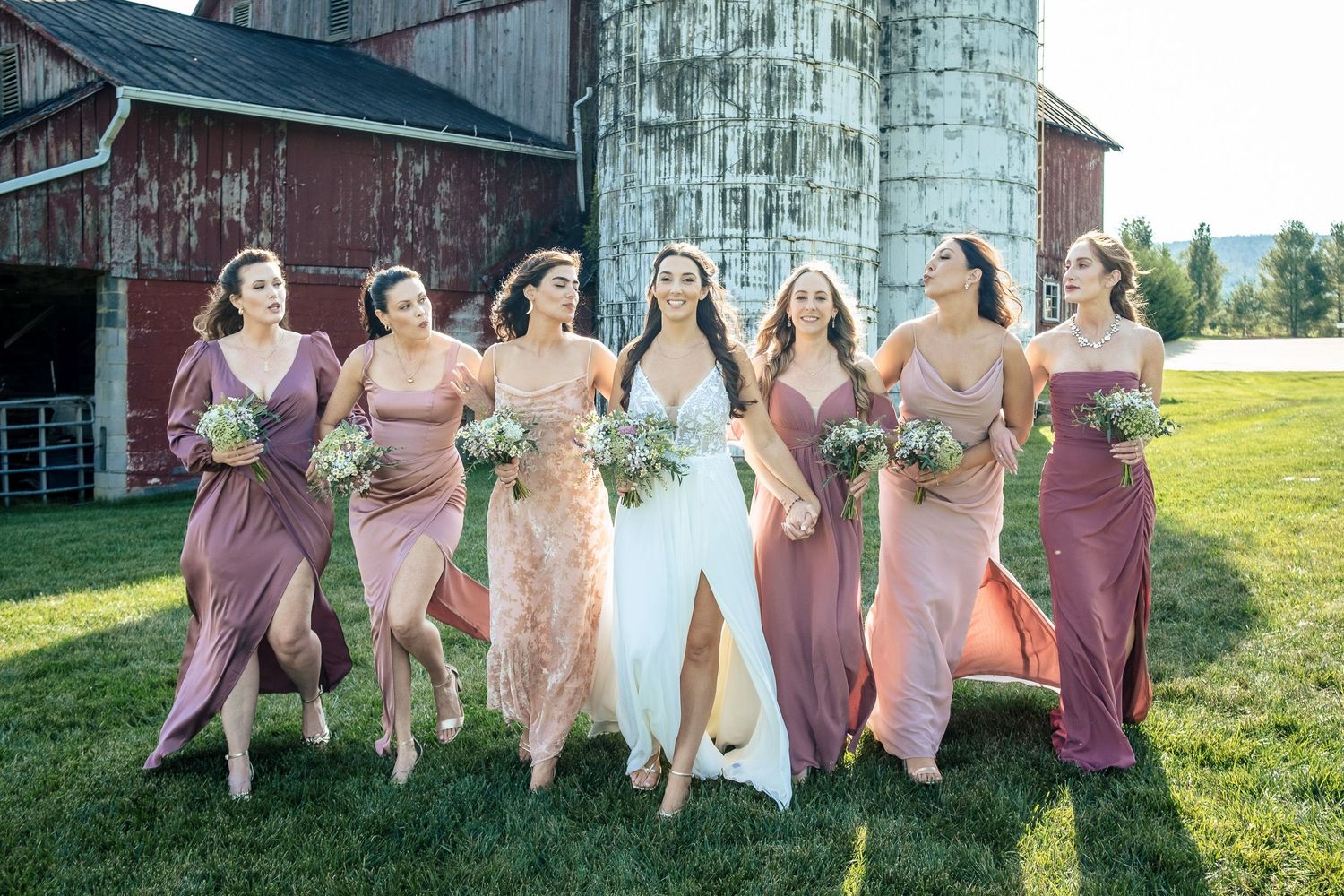Bride and bridesmaids in pink mismatched gowns walk together laughing near a red barn.