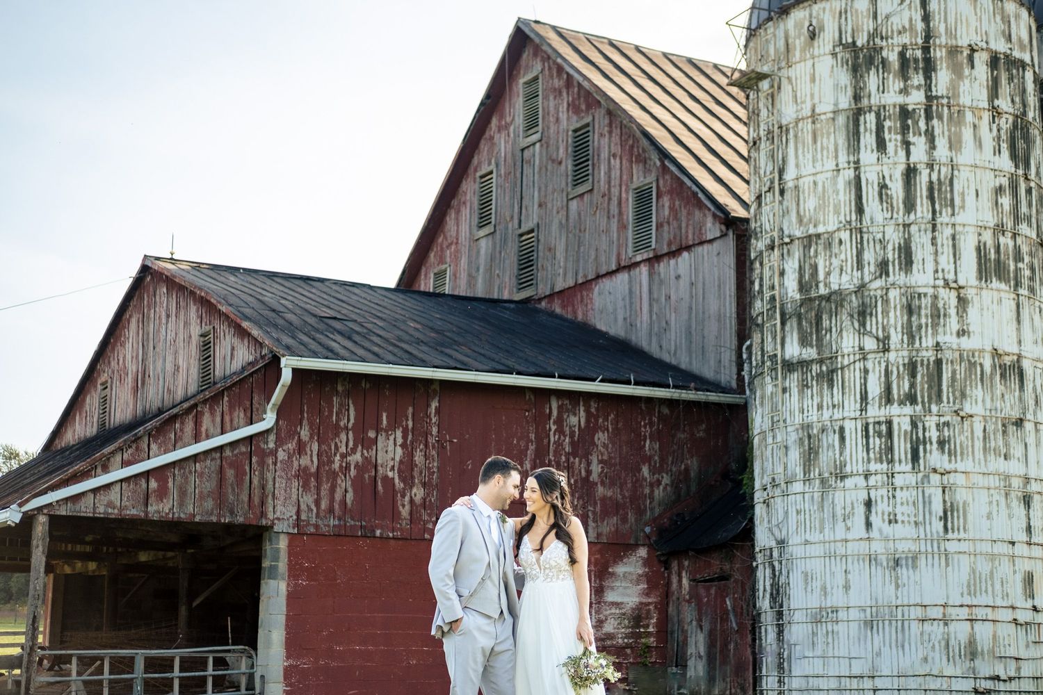 Bride and groom share a kiss beside a weathered stone silo and rustic red barn.