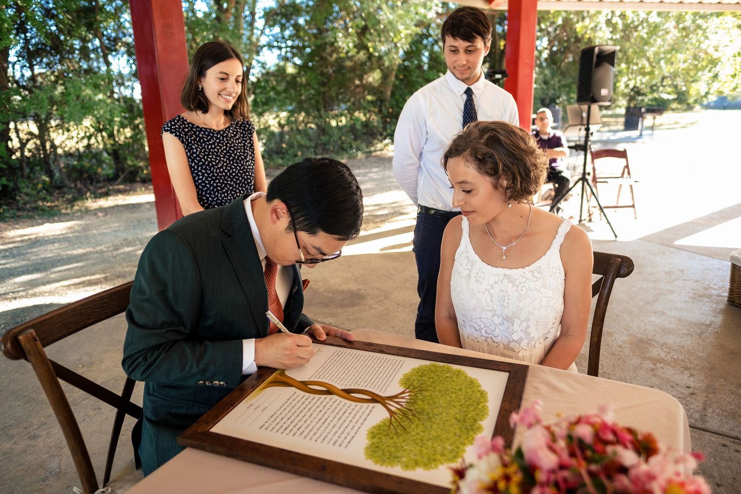 Groom signing wedding ketubah document at table with bride and witnesses watching outdoors.