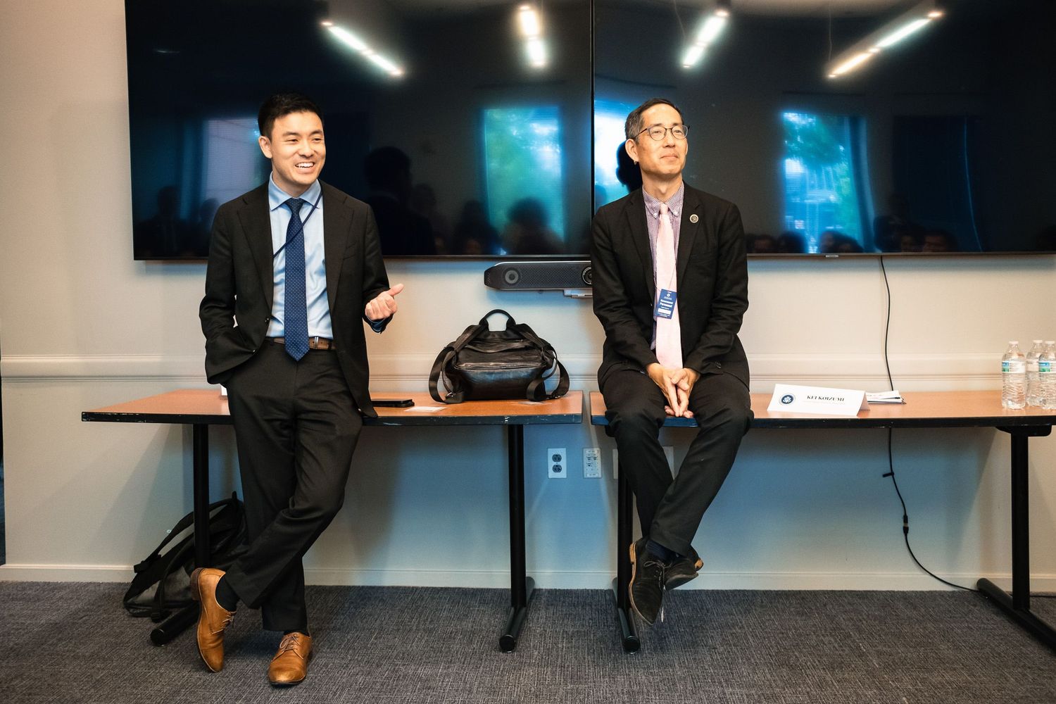 Two young men in suits sit casually on a table during a professional meeting or conference event.