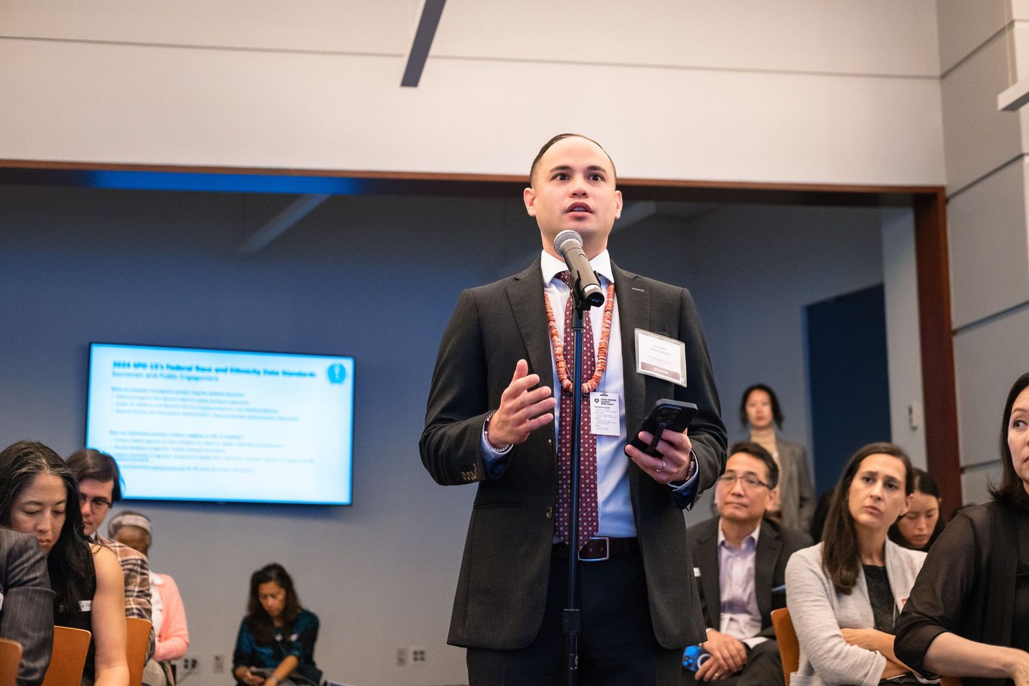 Professional speaker presenting at a business conference with audience members and a presentation screen in the background.