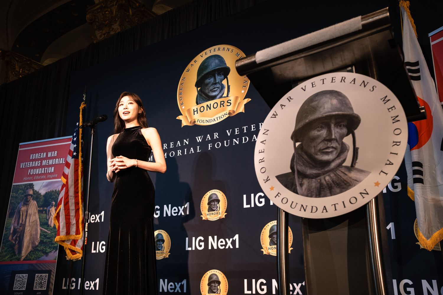 Elegantly dressed woman presents at Korean War Veterans Memorial Foundation gala with LIG Nex1 sponsorship backdrop.