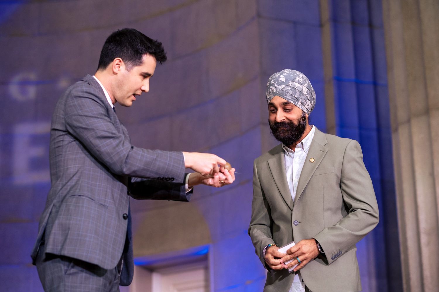 Two men in suits shaking hands on stage at a formal event, one wearing a blue turban.
