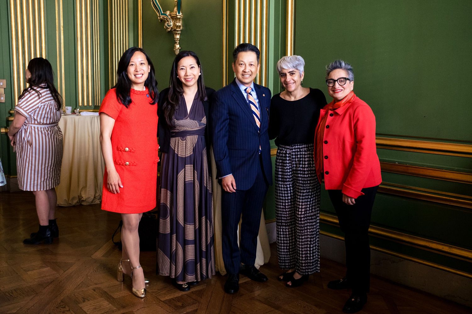 Five elegantly dressed people posing together in a green-walled formal venue at a special event.