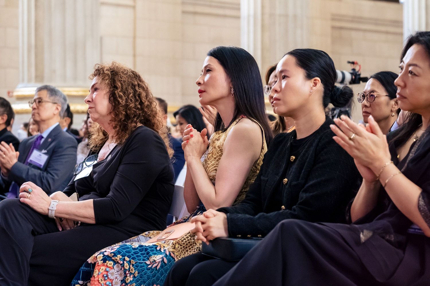 Diverse audience of elegantly dressed women seated at a formal indoor event, attentively watching a presentation.
