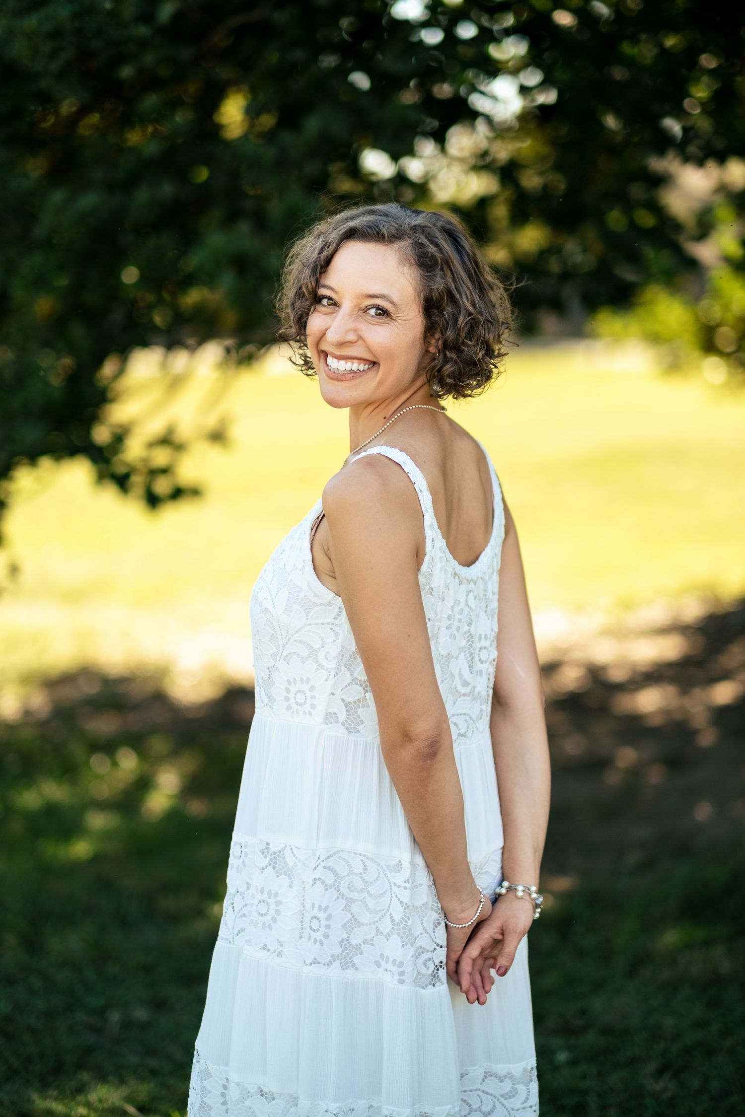 Smiling bride in a white lace dress glancing back over her shoulder in a sunlit green garden setting.