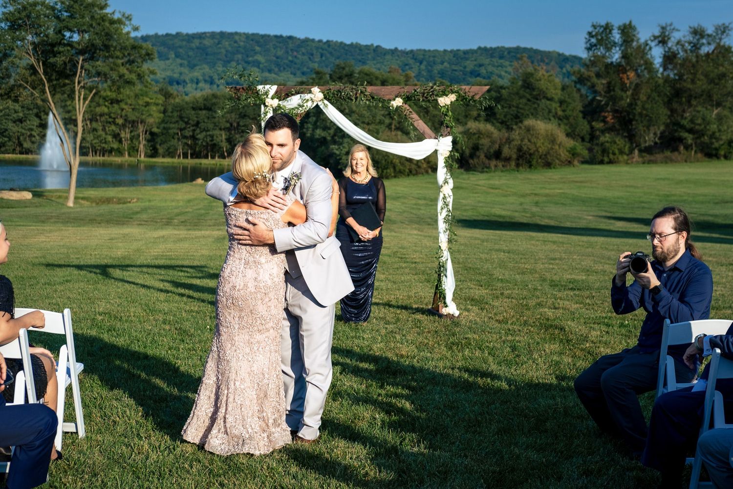 Groom embraces a family member near a floral wedding arch as a photographer captures the emotional moment.