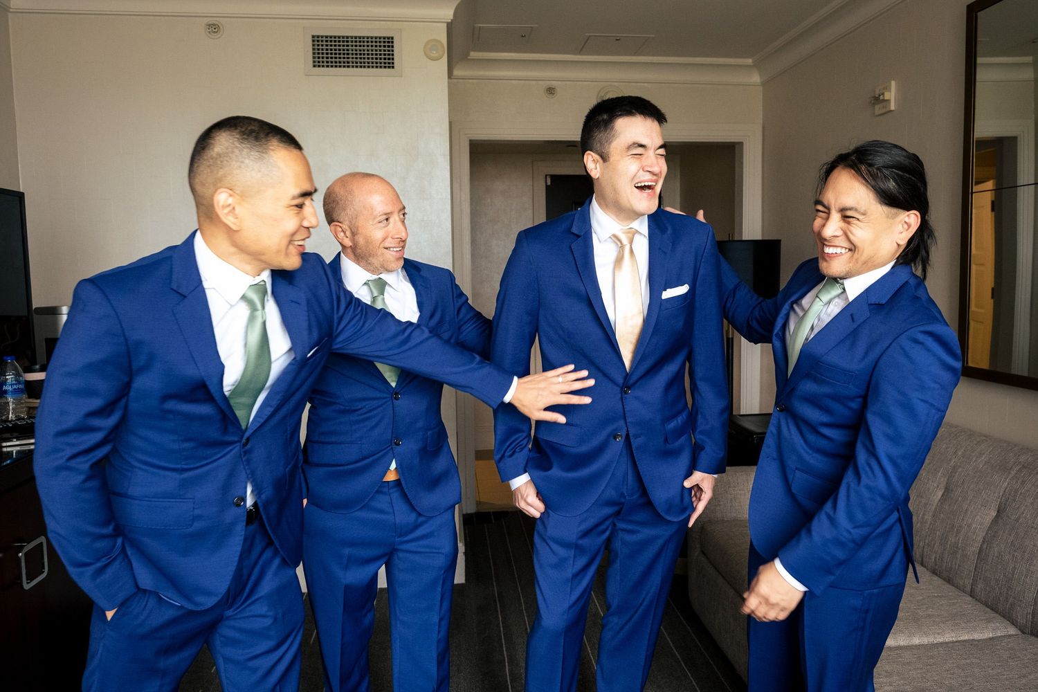 Four groomsmen in matching royal blue suits laughing together in hotel room before wedding.