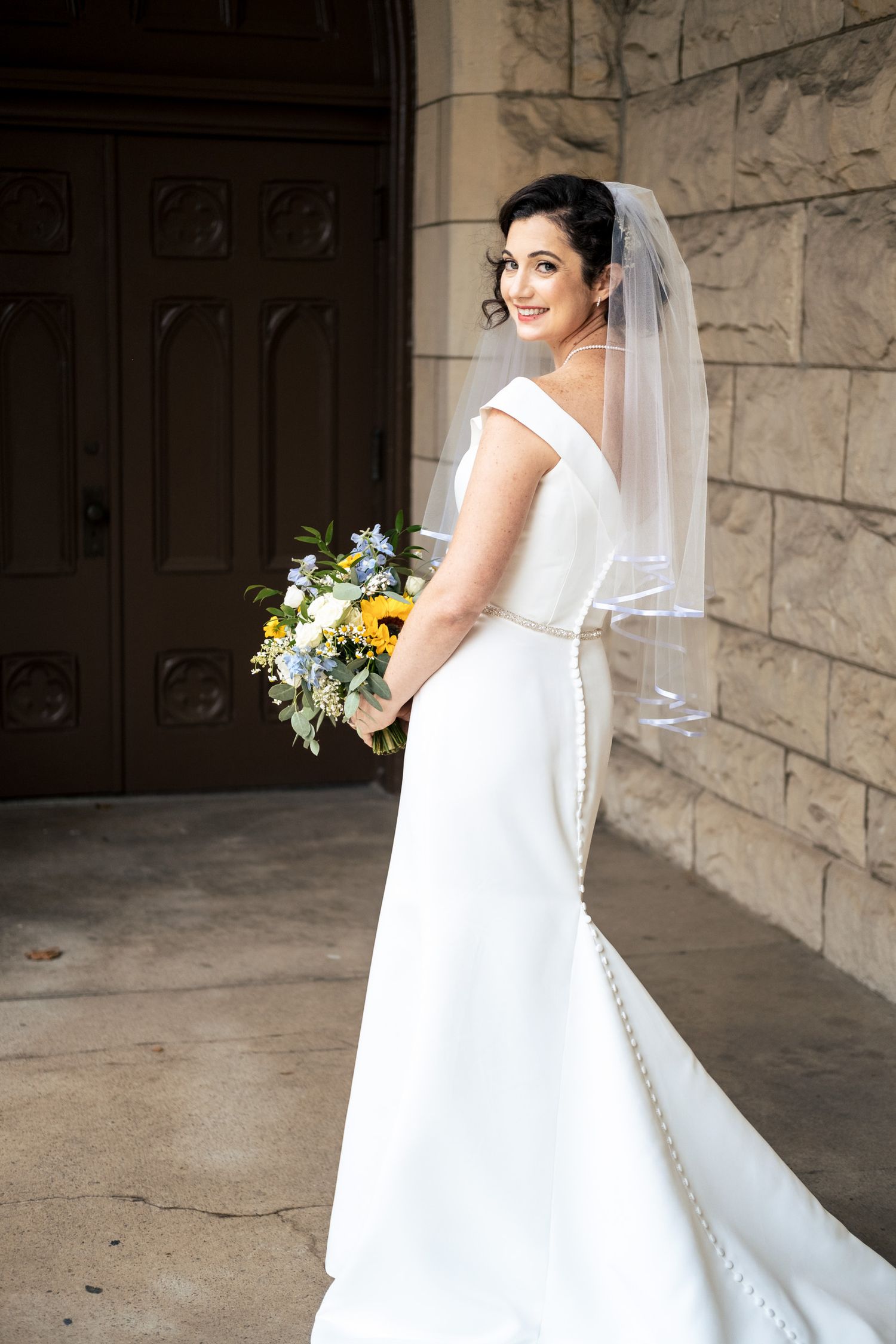 Bride in fitted white gown with button-back train poses with bouquet outside stone church entrance.