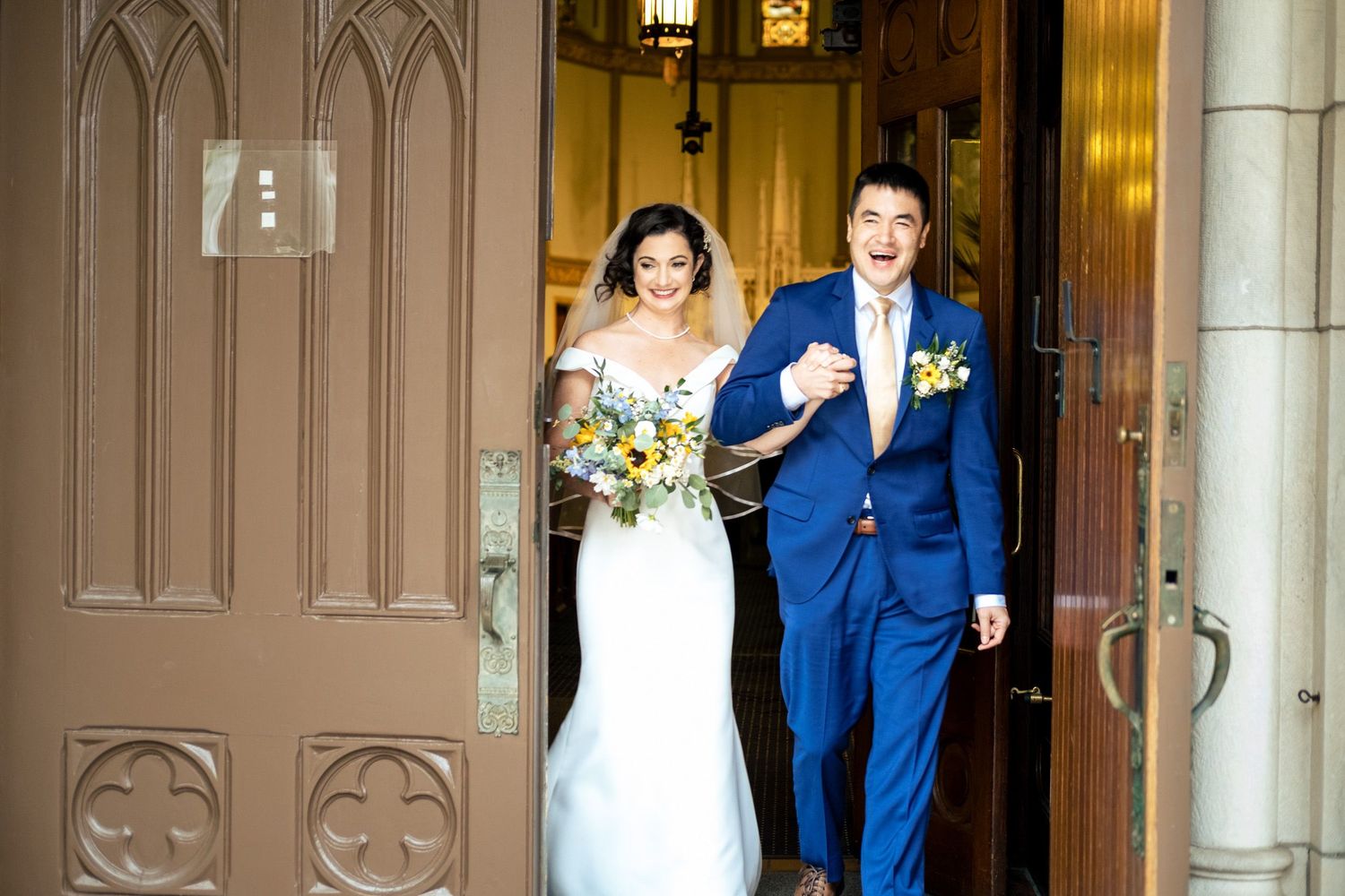 Newlywed couple exits through grand wooden church doors smiling after their wedding ceremony.
