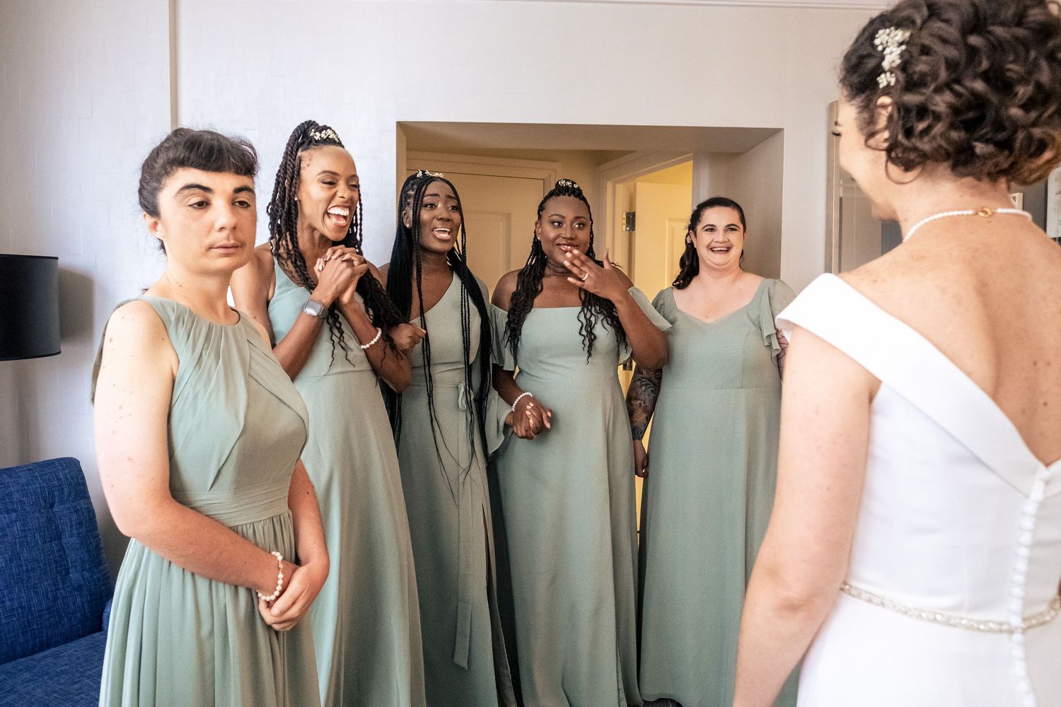 Four emotional bridesmaids in sage green dresses react with awe seeing bride for the first time.