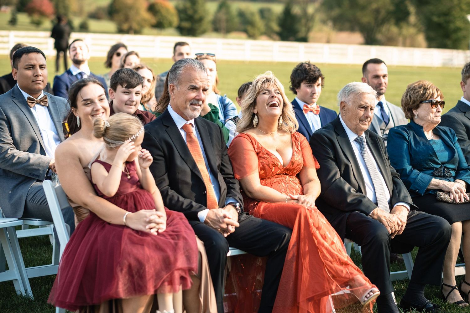 Wedding guests laugh and smile joyfully while seated at an outdoor ceremony, with a woman in orange standing out.