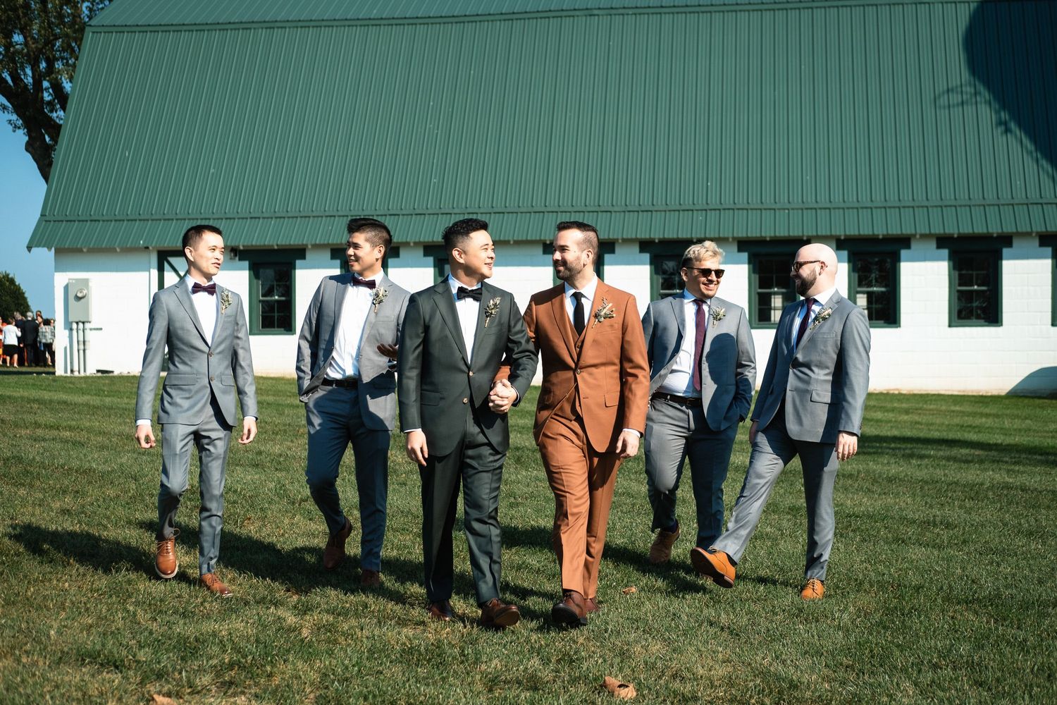 Two grooms and groomsmen walk together in coordinating suits on a sunny wedding day.