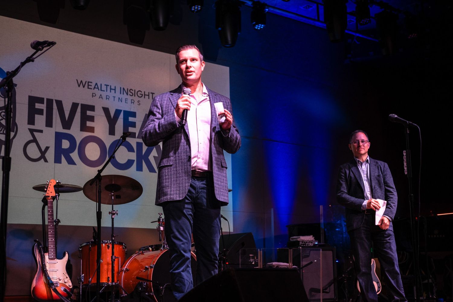 Two men speak on stage at Wealth Insight Partners Five Years & Rocking anniversary event with band instruments behind them.