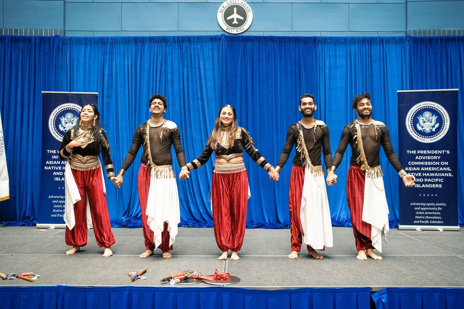 Five performers in traditional Indian dance costumes perform on stage with blue curtain backdrop.