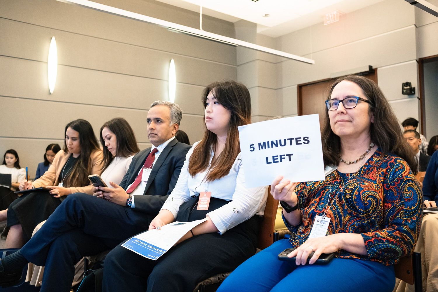 Audience members seated at a conference, with a woman holding a '5 Minutes Left' sign during a presentation.