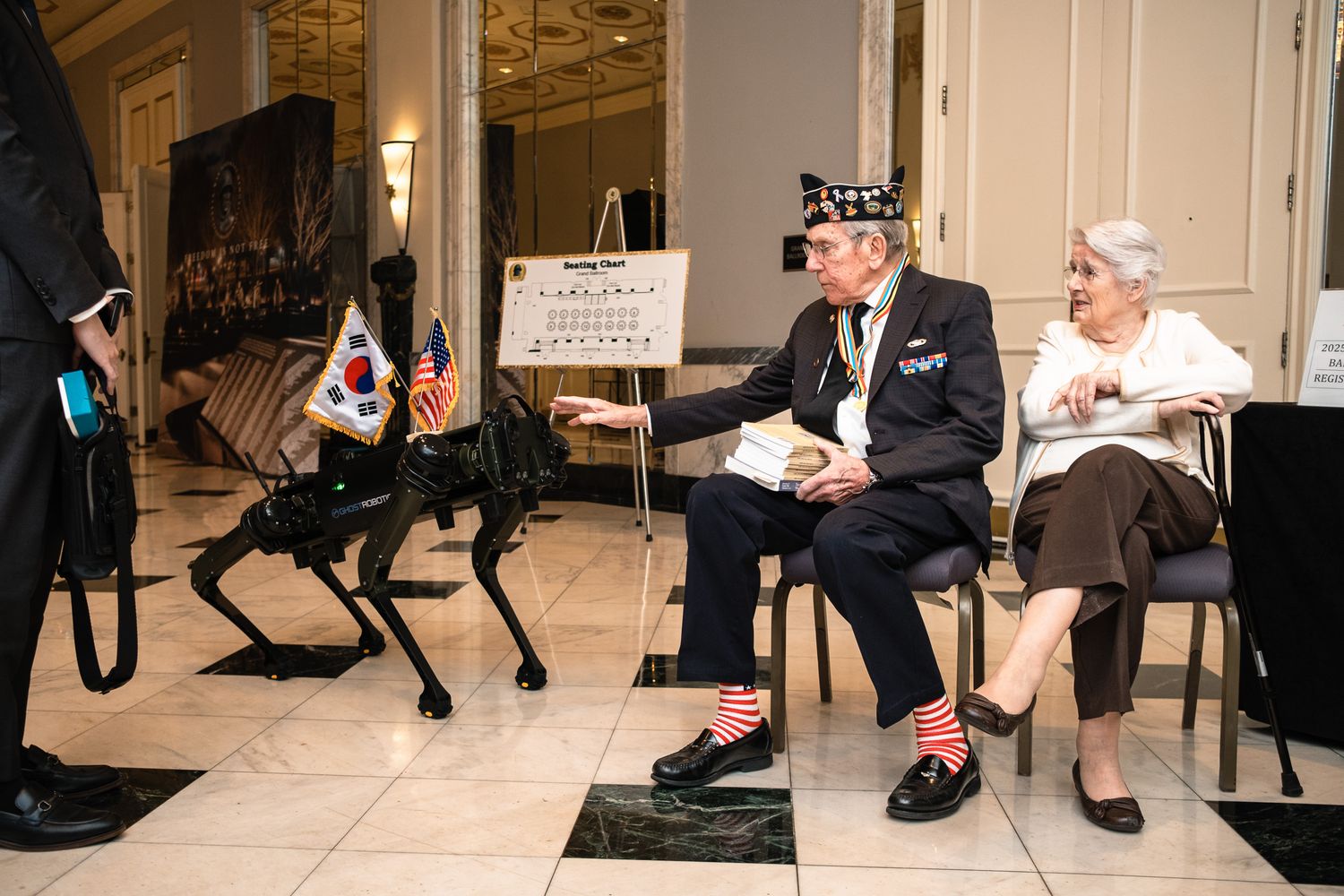 Veterans chat beside a robotic dog at a Korean War memorial event with US and South Korean flags displayed.