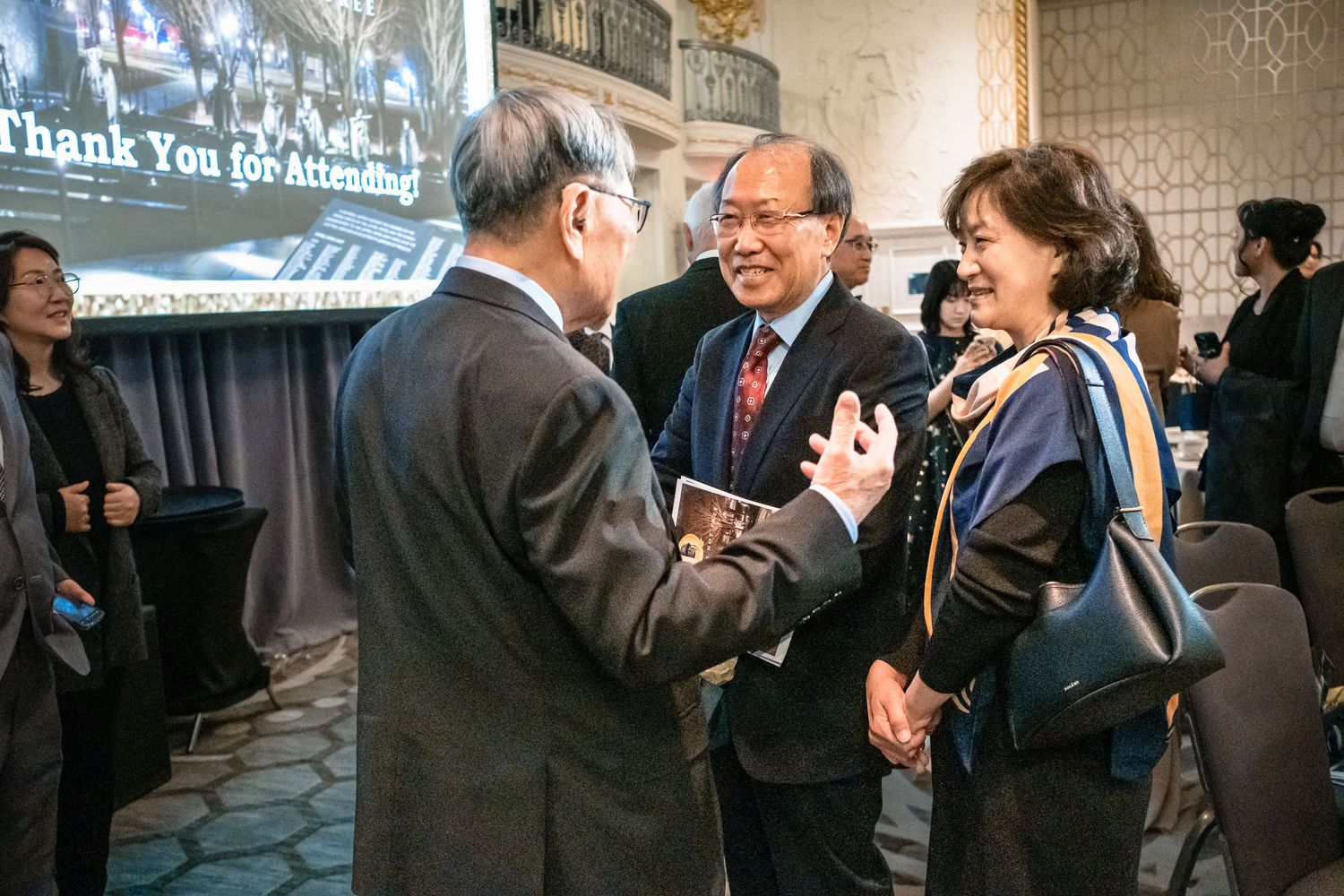 Three attendees engage in conversation at a formal event with a Thank You for Attending screen.