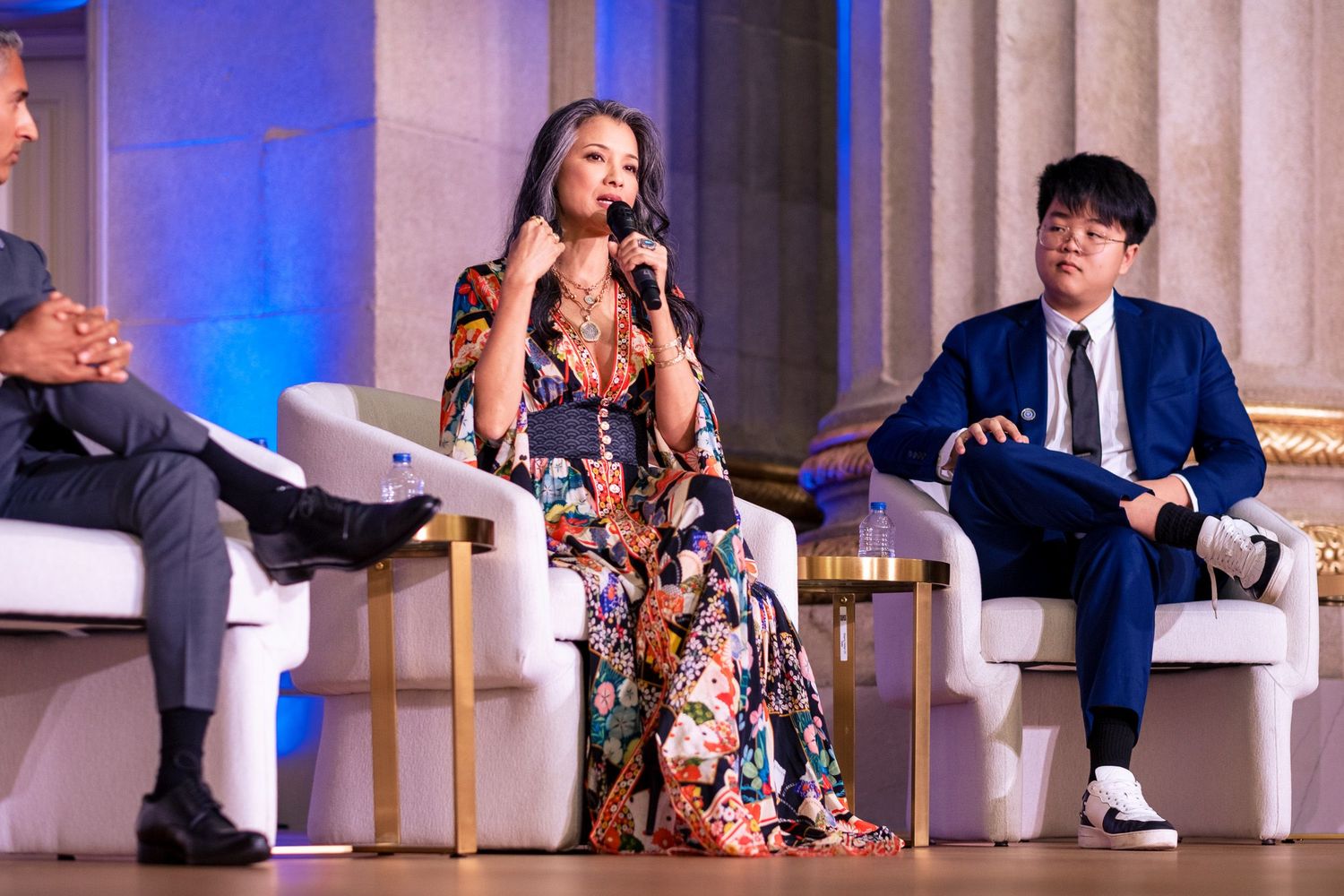 Three panelists on stage at a formal event, woman in floral dress speaking into microphone flanked by two men.